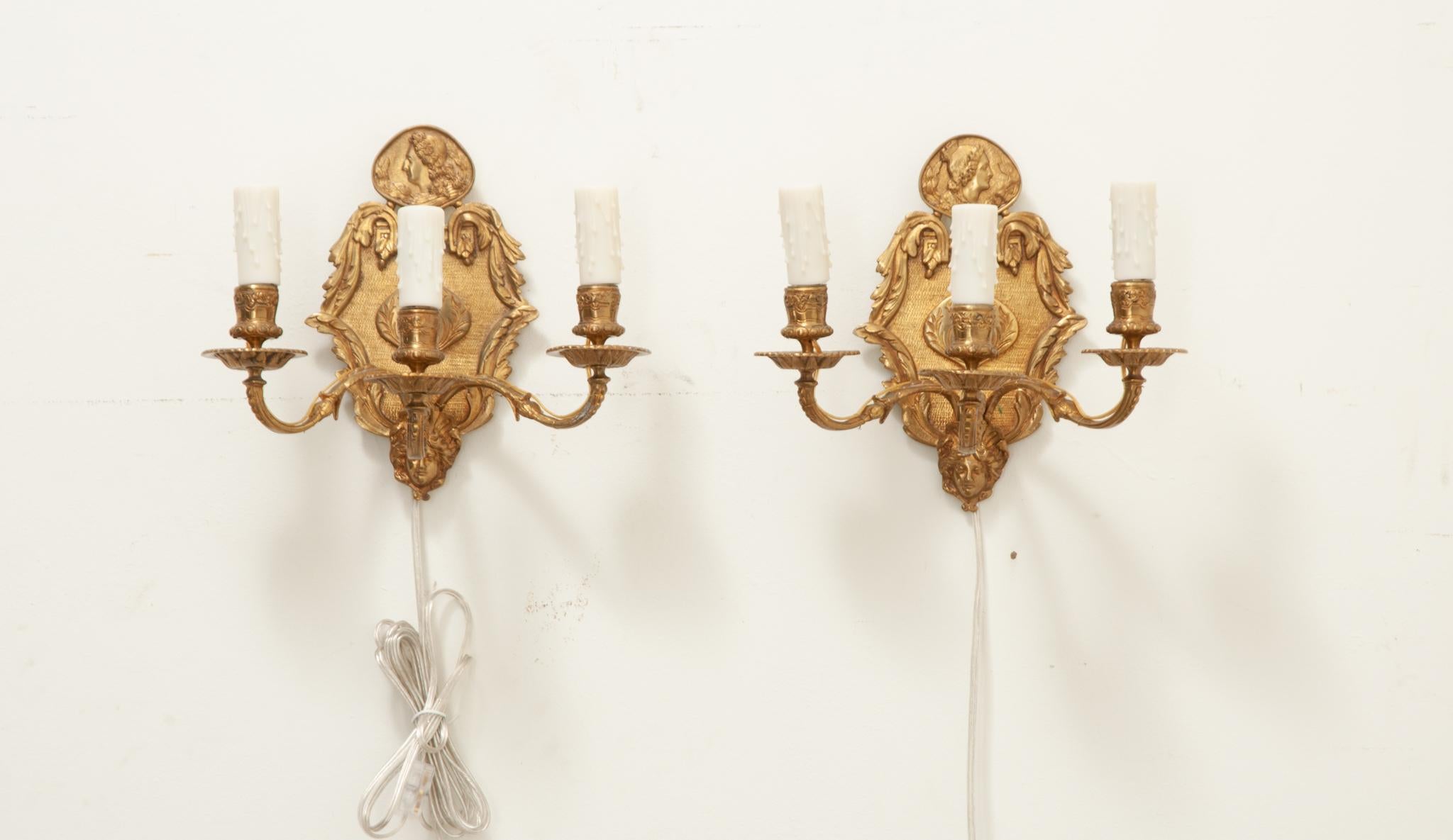 An elegant pair of French brass wall sconces. This pair of petite sconces has three brass arms each with faux candle covers. Classic French motifs are throughout the fixture, including a crest of fleur de lis topped with a crown and pendants