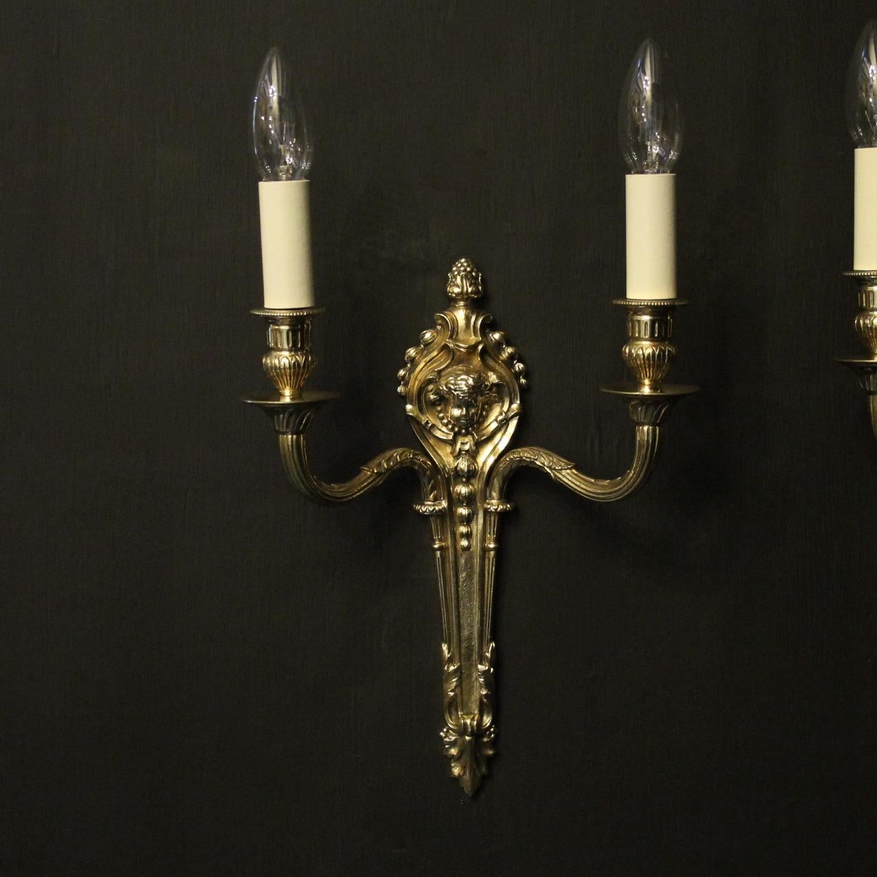 A French pair of repolished brass twin branch antique wall lights, the reeded leaf scrolling arms with circular twisted bobeche drip pans and leaf bulbous candle sconces, issuing from a tapering backplate with cherub headed centre and berried