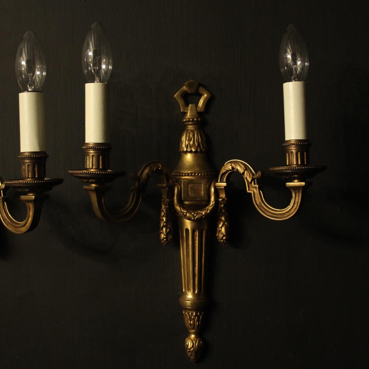 A French pair of gilded bronze twin branch antique wall lights, the leaf clad reeded scrolling arms with circular bobeche drip pans and millgrain reeded candle sconces, issuing from a laurel swaged foliate reeded tapering back plate, with pierced