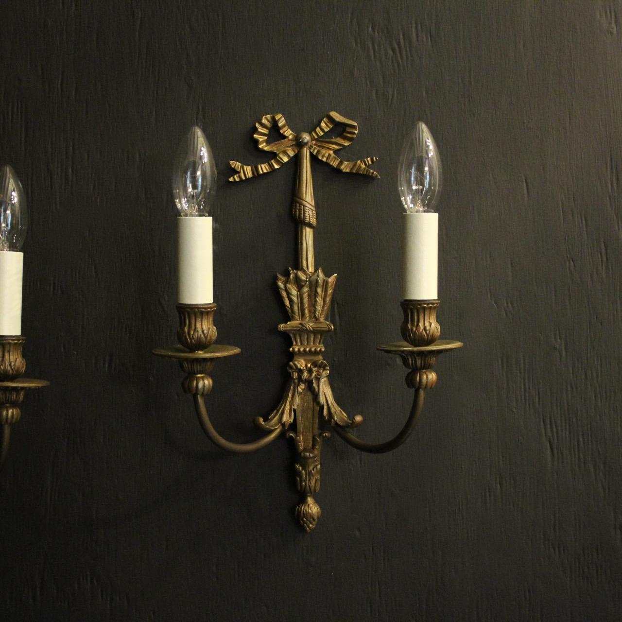 A French pair of gilded bronze twin arm antique wall lights, the scrolling arms with leaf bobeche drip pans and reeded candle sconces, issuing from an ornate tapering arrow sheath back plate with pierced ribbon finial, nice proportions with good