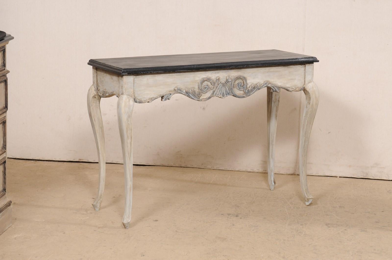 A French pair of carved and painted wood console tables. These vintage tables from France each have a slightly overhanging rectangular-shaped top, with molded edging, over scalloped aprons which are embellished with a carved shell flanked within