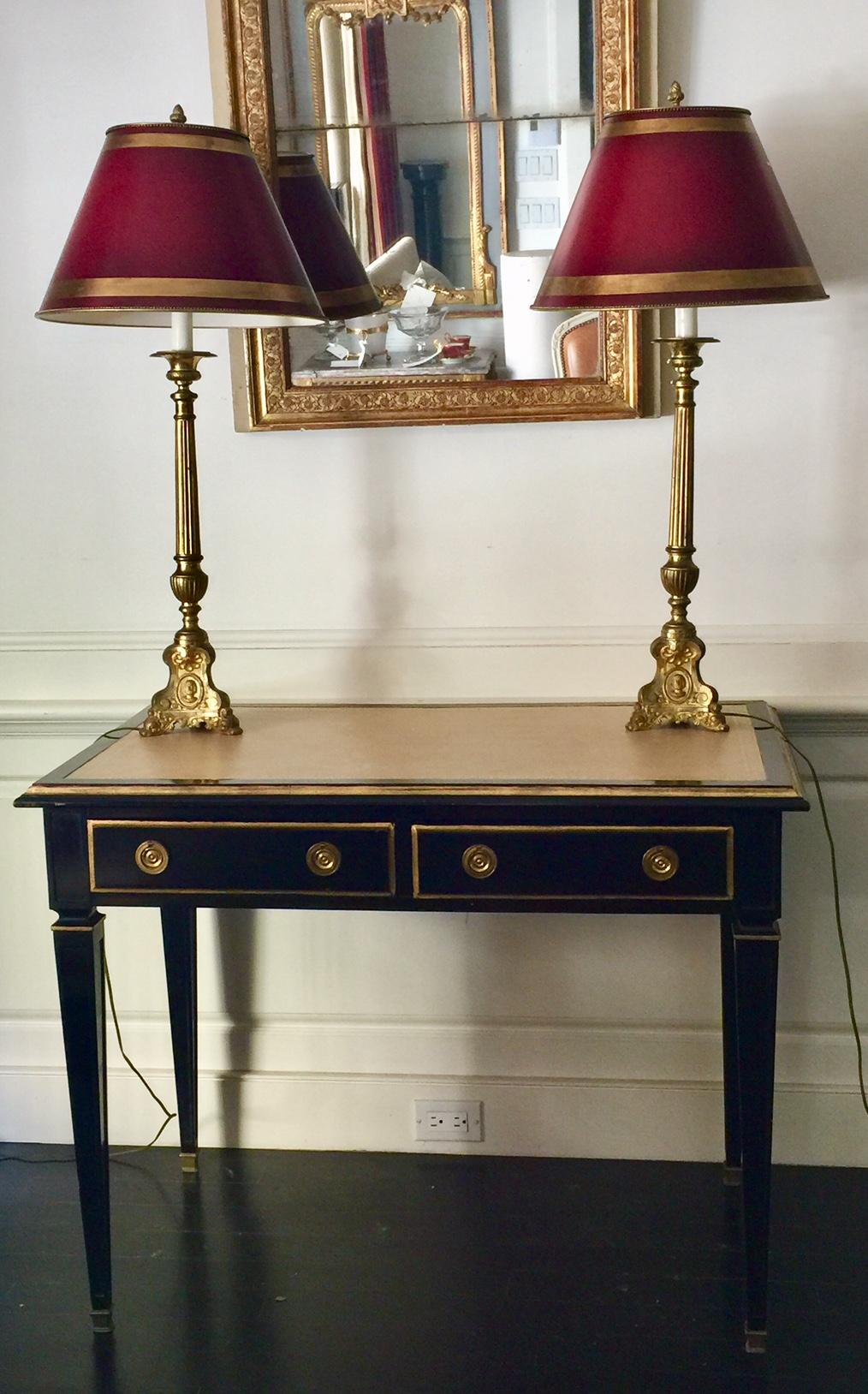 French pair of column lamps with burgundy red parchment shades.
Tall and highly decorative pair of column lamps with burgundy red shades trimmed in wide gold bands. The columns rise from the tripod base decorated with escutcheons, to the acorn