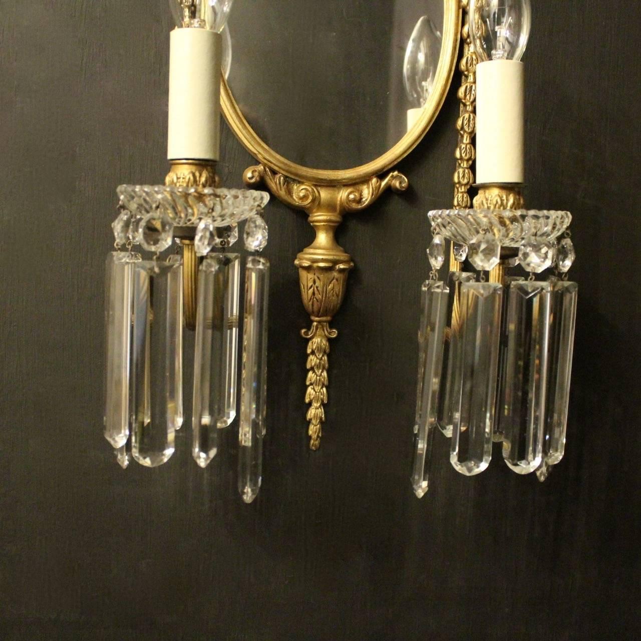 A French pair of gilded cast bronze twin arm antique girandoles, the reeded scrolling arms with glass bobeche drip pans and bulbous leaf candle sconces, issuing from an ornately cast oval mirrored backplate with central floral basket final and