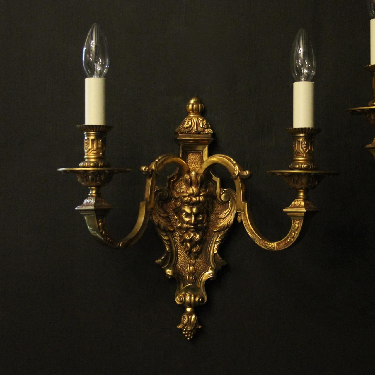 A French pair of gilded bronze Bacchu headed twin arm antique wall lights, the leaf clad scrolling arms with sectional leaf bobeche drip pans and ornate candle sconces, issuing from a decoratively cast backplate with Bacchus headed central mask,