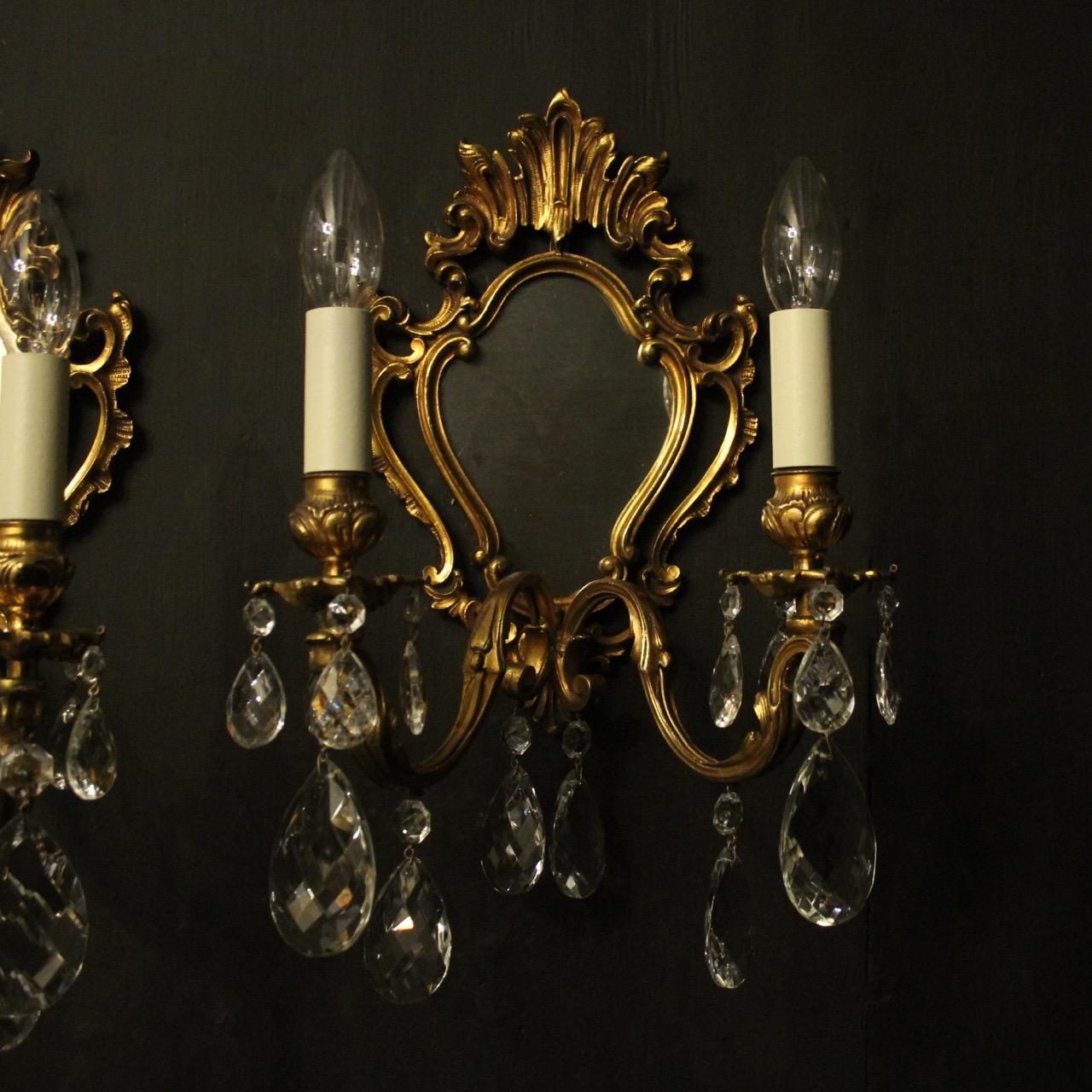A French pair of gilded cast bronze twin arm antique girandoles, the ornate leaf scrolling arms with foliated bobeche drip pans and bulbous candle sconces, issuing from an ornately cast cartouche leaf mirrored backplate and decorated with faceted