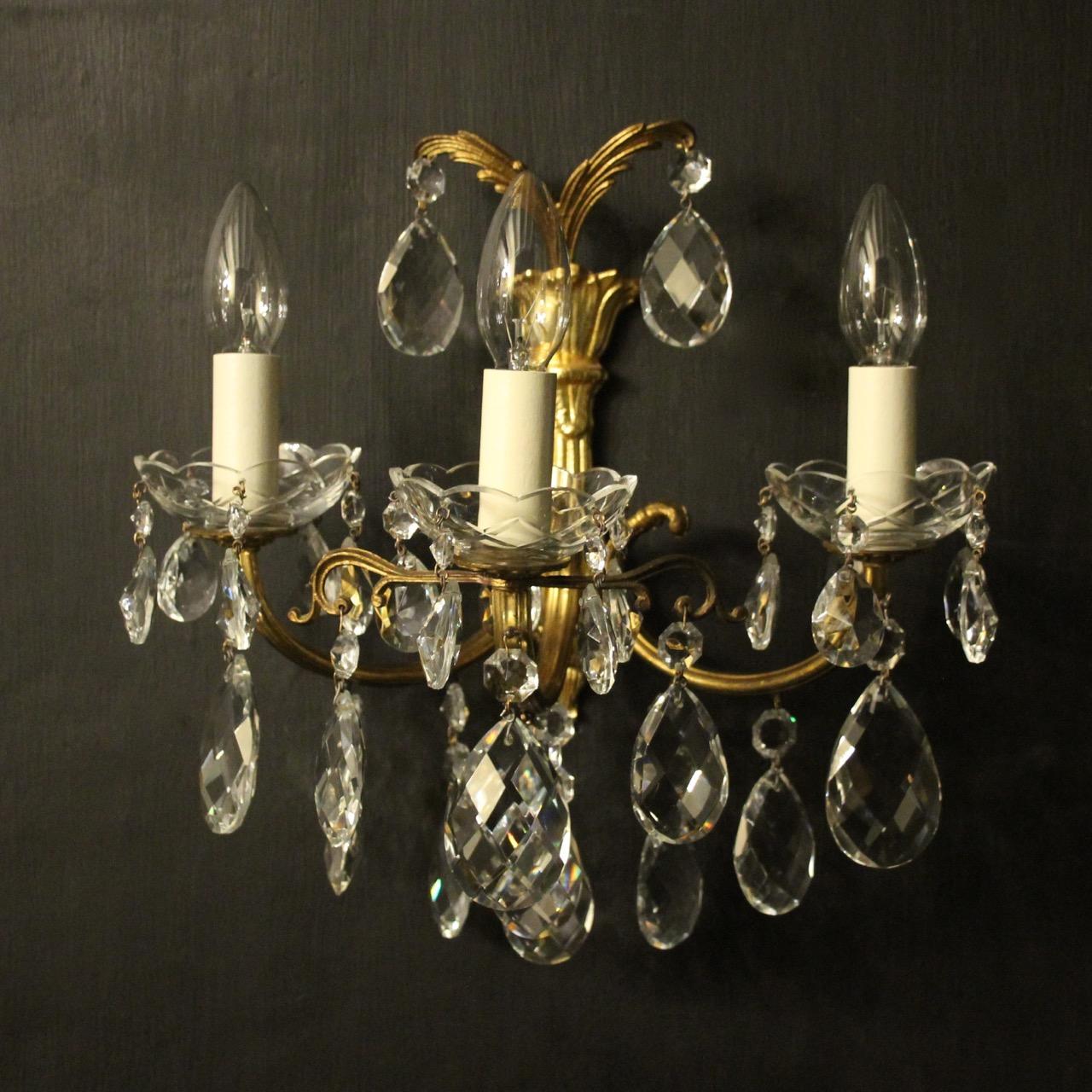 A French pair of gilded brass and crystal triple arm antique wall lights, the leaf scrolling arms with glass bobeche drip pans, issuing from a foliated leaf backplate with twin leaf canopy, decorated overall with crystal faceted pendants. Fully