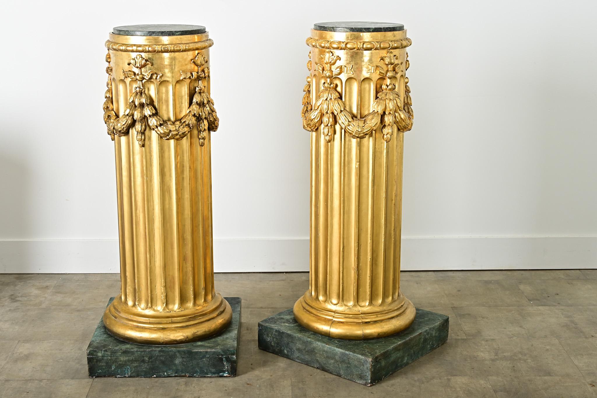 A bold pair of French 19th century giltwood columns for Neoclassical beauty in any interior. Each radiant column is topped with a disc of green marble to elegantly elevate a plant or vase, accented with festoons wrapping the circumference that drape