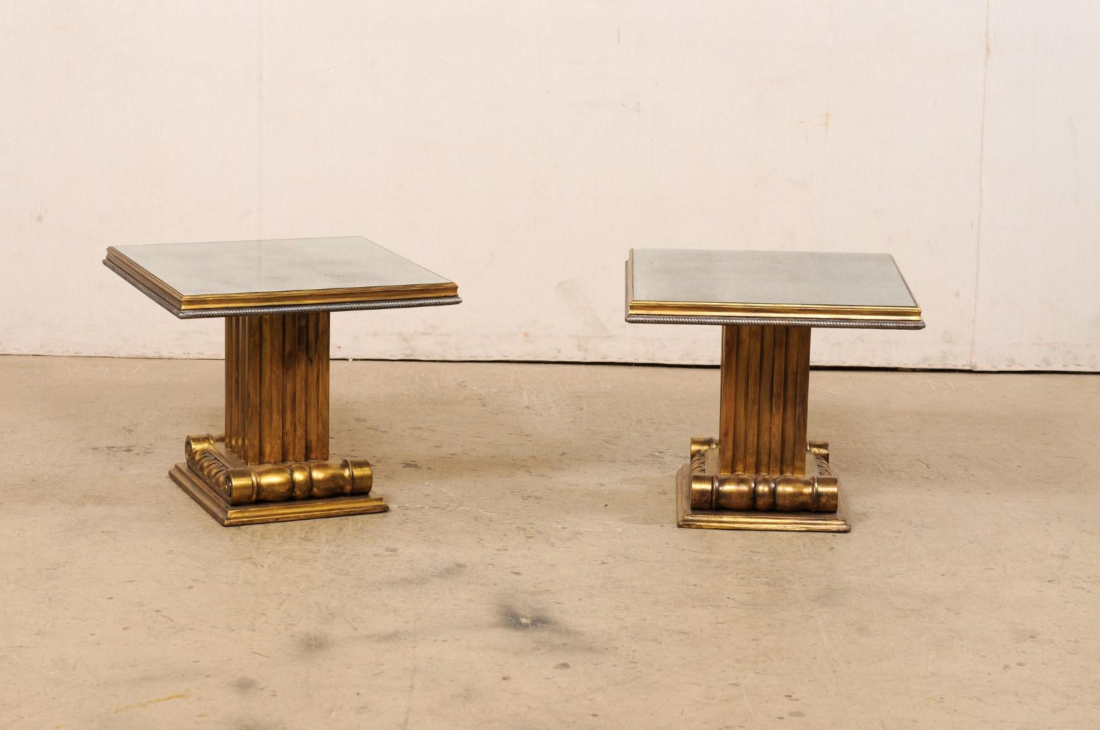 A French pair of gold & silver column side tables with antiqued mirror tops. These vintage tables from France each feature square-shaped tops, recessed with antiqued mirrors set within a molded lip with roped trim about the bottom edge, and