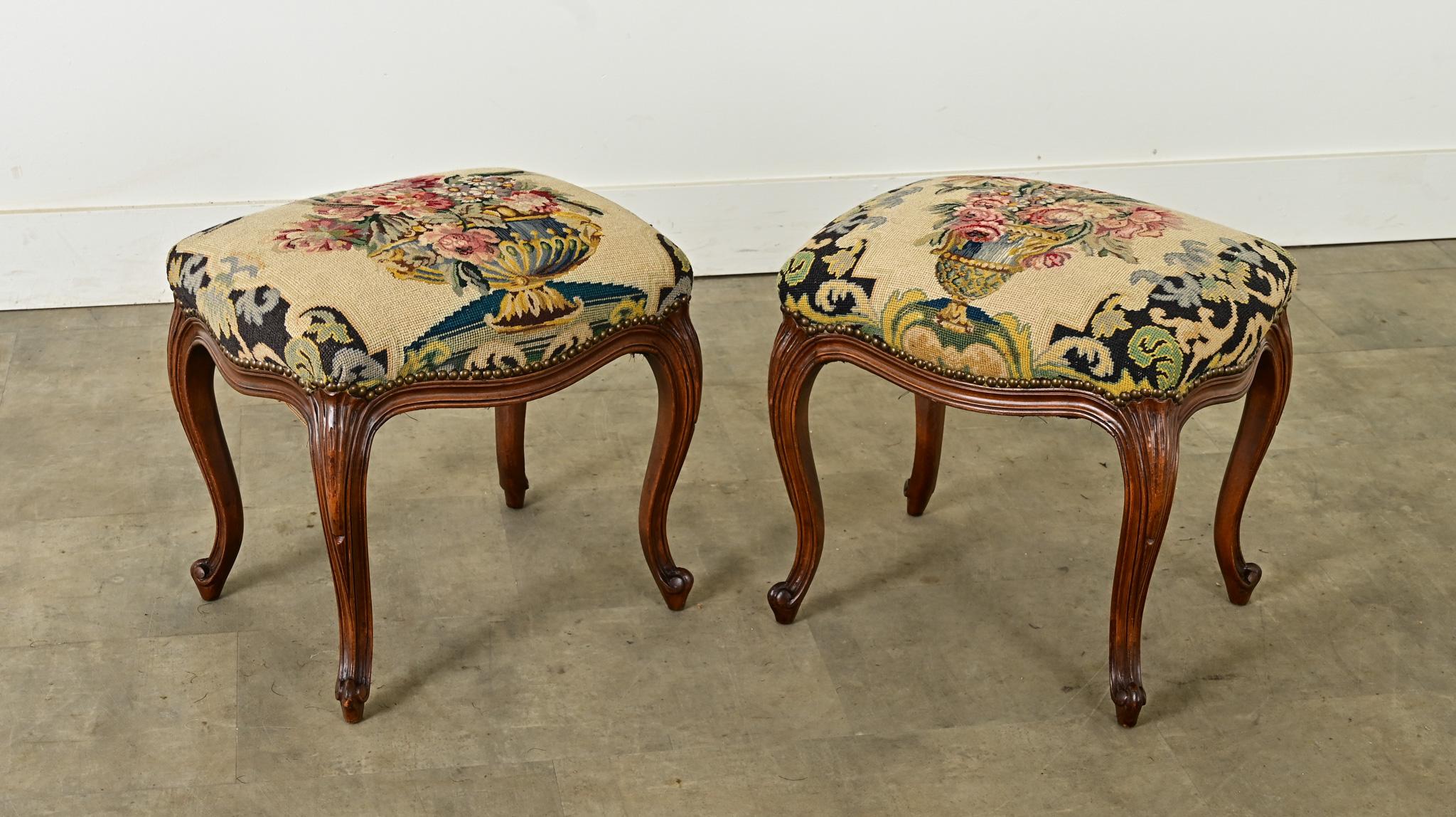 Paire de poufs de style Louis XV avec tapisserie à l'aiguille fleurie. Chaque tabouret présente un design unique mais coordonné d'urnes ornées débordant de bouquets de fleurs colorés et luxuriants sur fond beige, avec une finition en tête de clou.