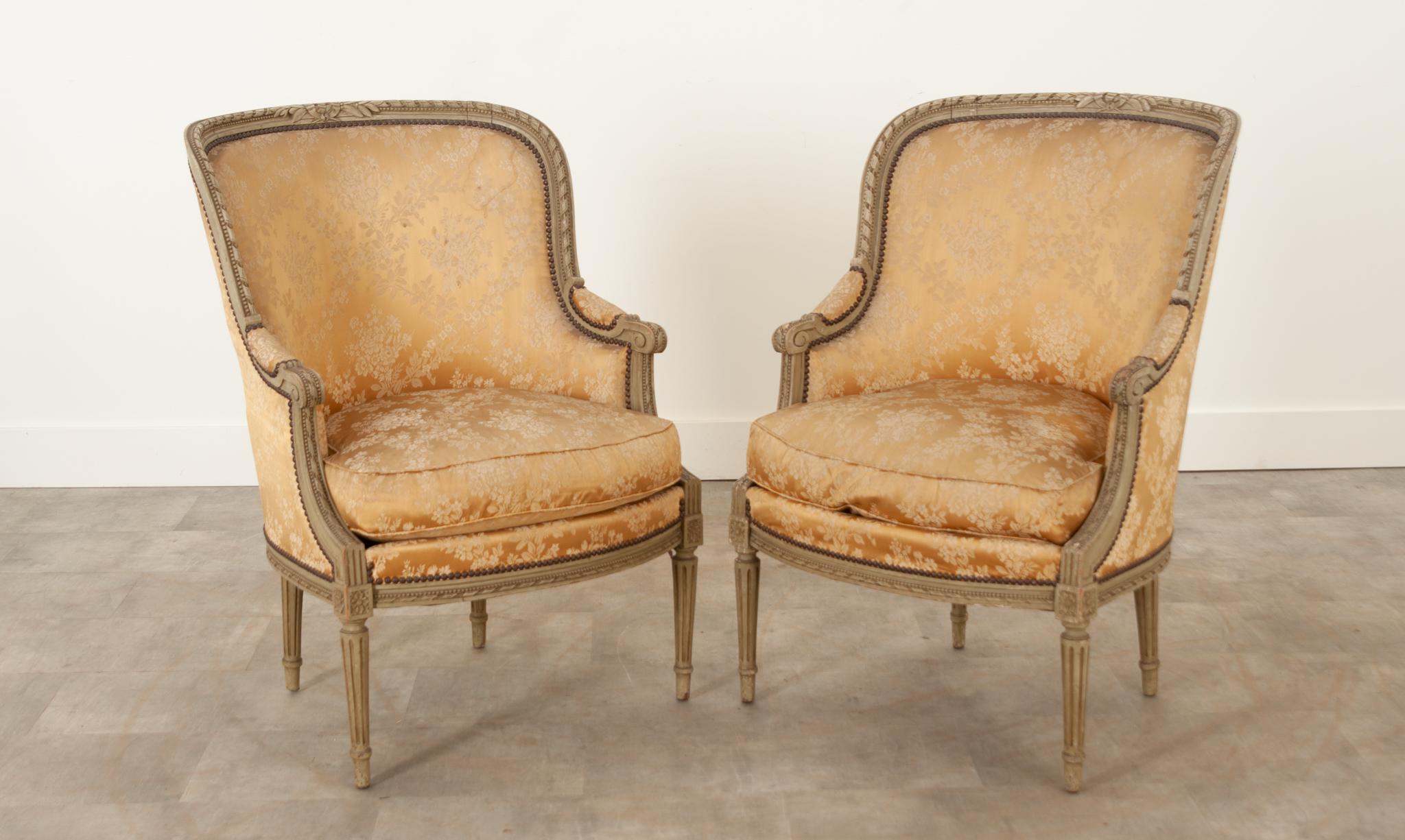 This pair of French Louis XVI-Style bergeres are in wonderful antique condition. The frames are 19th century, hand carved with a twisted ribbon border and painted in its original worn gray finish. Upholstered in elegant champagne toned, silk floral