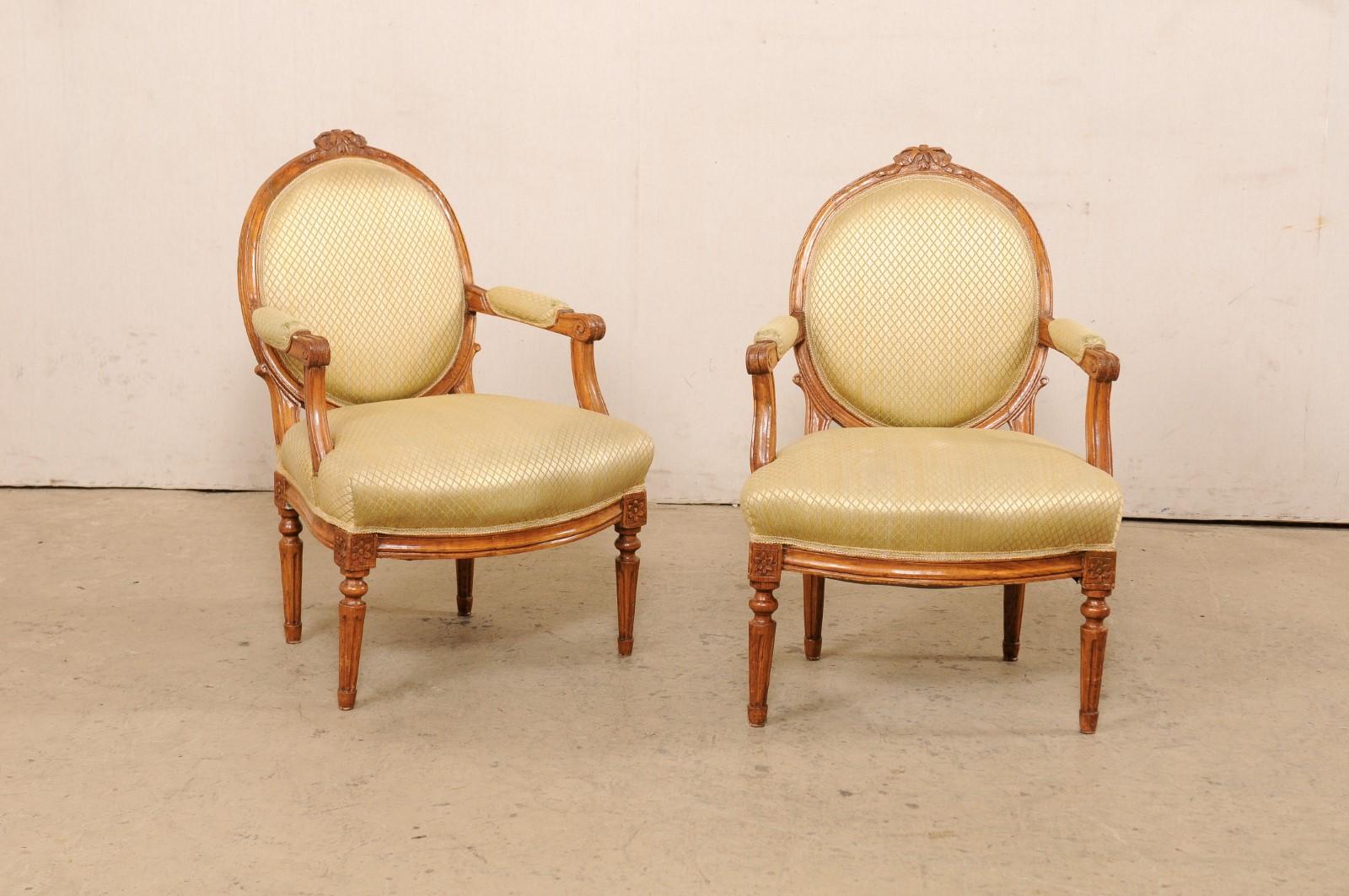 A French pair of Louis XVI style carved wood and upholstered fauteuil chairs from the mid 20th century. This mid-century pair of armchairs from France each feature oval-shaped upholstered backs within a wood frame accentuated with spiraled ribbon
