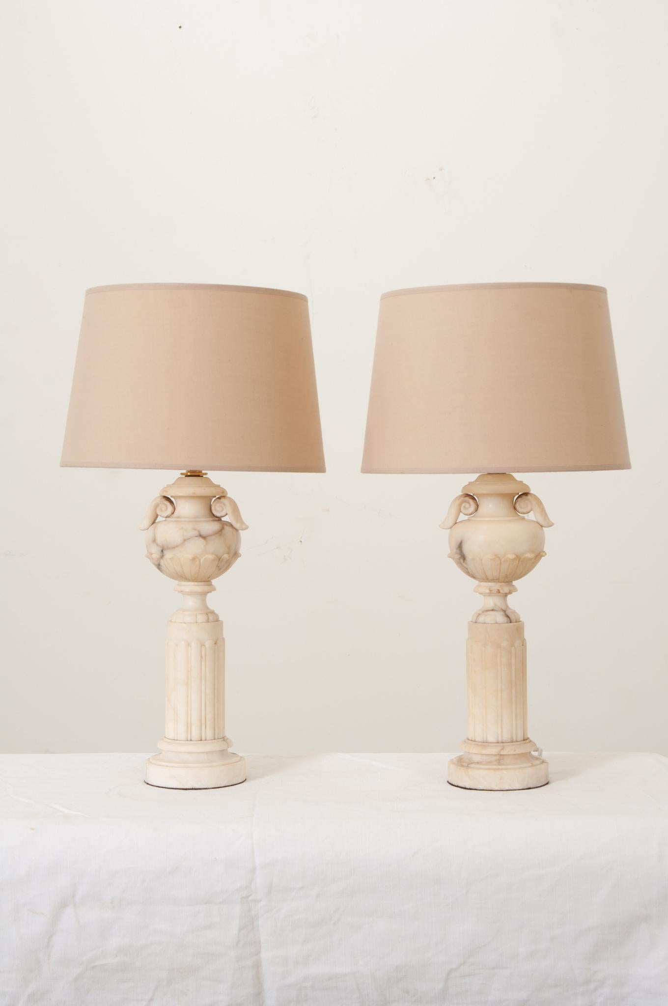 A French pair of carved white marble column lamps with urns. Elegant in style, with years of patina, these lamps can go in any interior. European mount shades complement this pair of table lamps. Cleaned and rewired for US electrical, ready for your