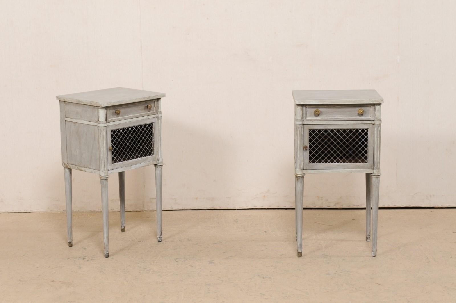 A French pair of neoclassic style painted wood side chests with wire front doors. This vintage pair of end tables from France each have a nearly-square shaped top which rests above a house which is fitted with a small drawer at top, over latticed