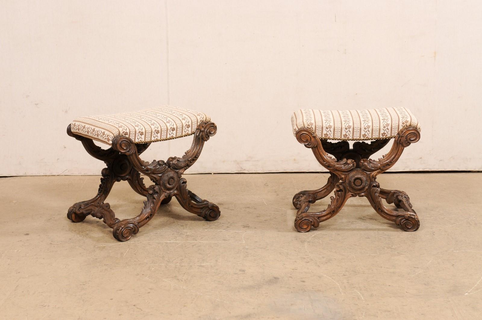 A French pair of ornately carved wood and upholstered stools from the 19th century. These antique stools from France each have rectangular-shaped upholstered seats which are presented upon elaborately x-style wood frames carved in a motif of