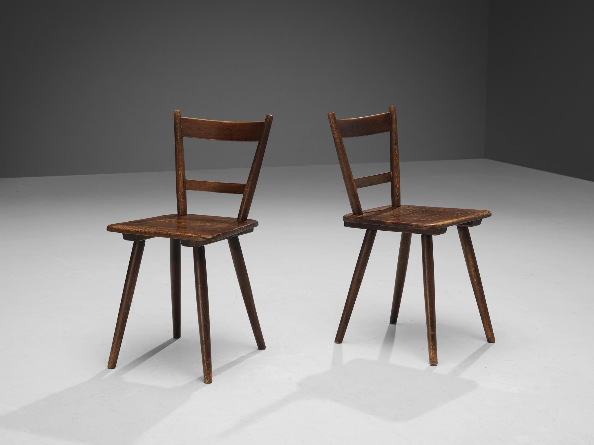 Pair of dining chairs, stained wood, France, late 1940s. 

Rustic pair of dining chairs manufactured in France. Simple yet stylish, these chairs feature a modest frame with sleek, elegant elements. Note for example the tapered legs slightly pointing