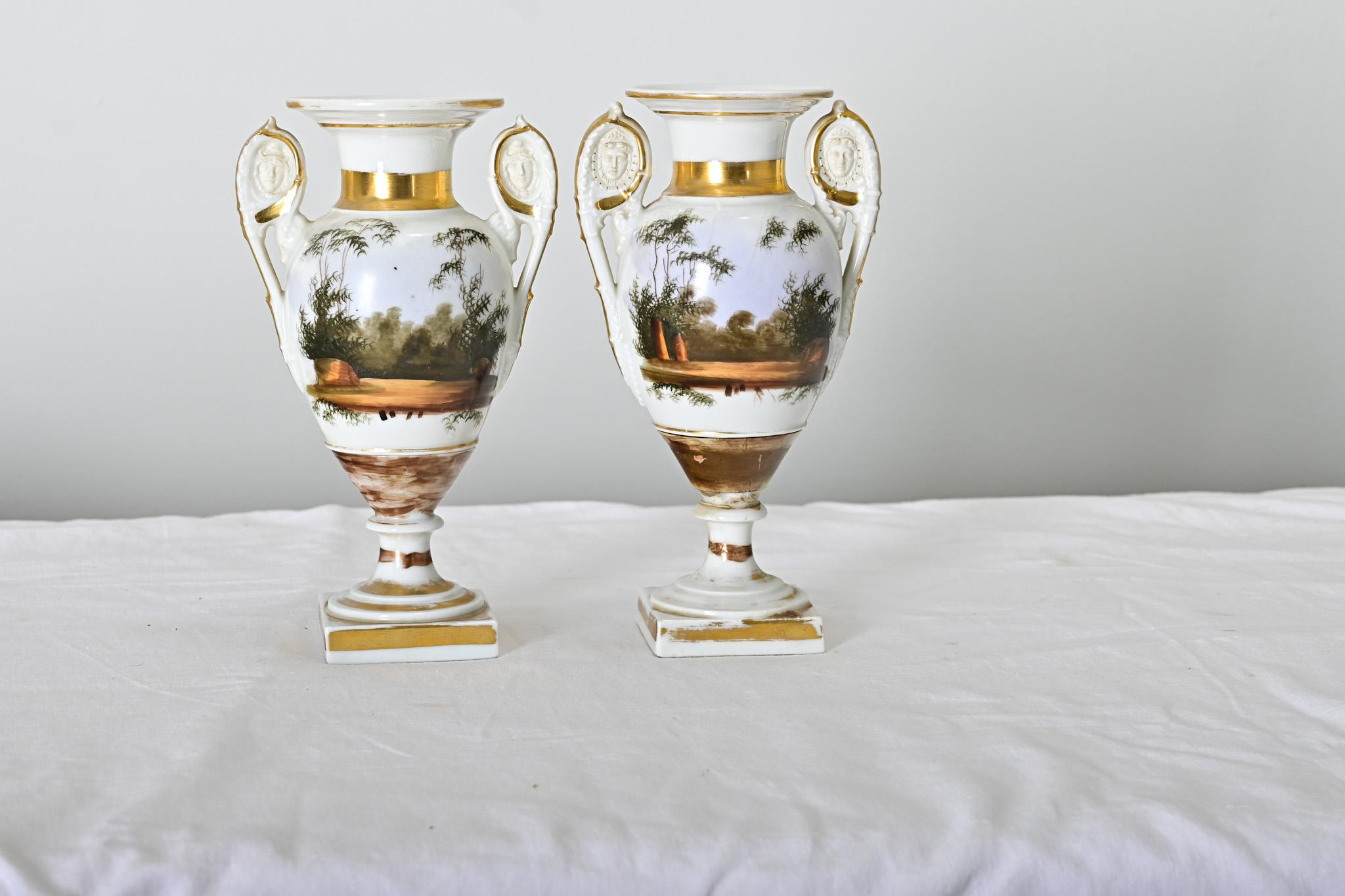 Coppia di vasi in porcellana Old Paris del periodo Impero francese, 1810 circa, con paesaggi dipinti e scene figurative su ciascun lato. Il profilo classico dell'anfora è caratterizzato da una bocca bordata d'oro nella parte superiore, che passa al