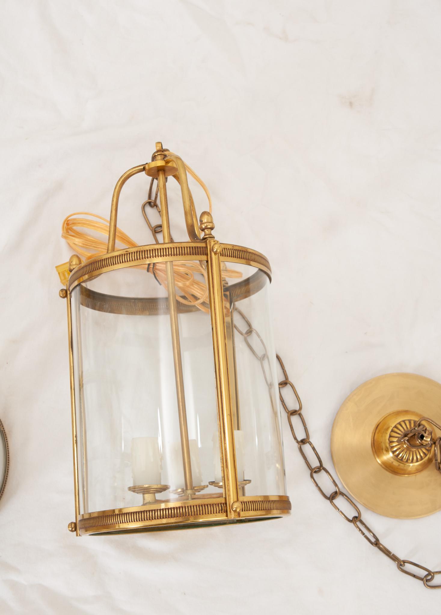A Pair of petite French brass lanterns are simple in design and have curved glass panels. Each lantern has three candle cups. Recently wired for US electrical using UL listed parts, ready to be hung in your interior. 3’ of chain, 6 ¼” diameter