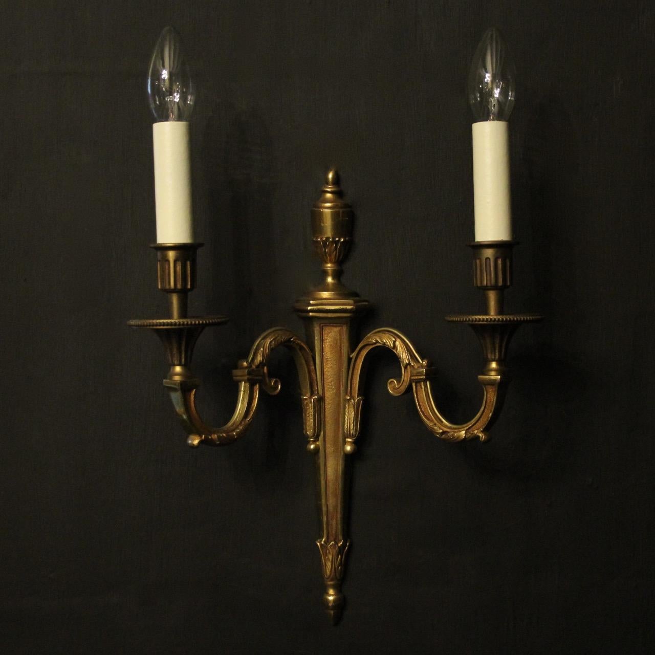 A French pair of gilded cast brass twin arm antique wall lights, the tapering square gauge leaf scrolling arms with trumpet reeded bobéches mill grain drip pans and reeded candle sconces, issuing from a decoratively cast elongated tapering backplate