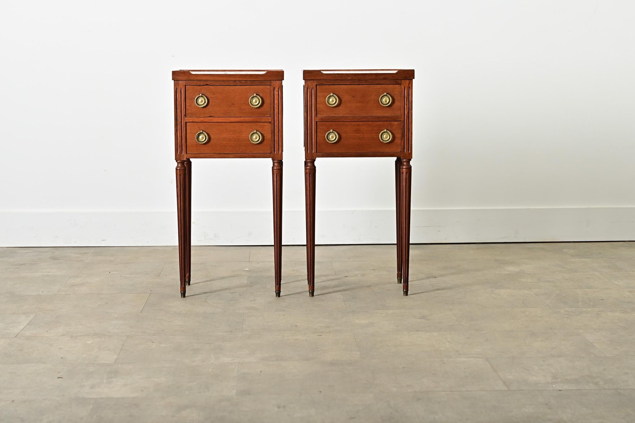 Paire de petites tables de chevet françaises du milieu du 20e siècle offrant deux tiroirs dans des cadres en acajou. Les plateaux en marbre blanc encastrés sont entourés d'un court bord de galerie et coupés par une ouverture basse à l'avant. Cet