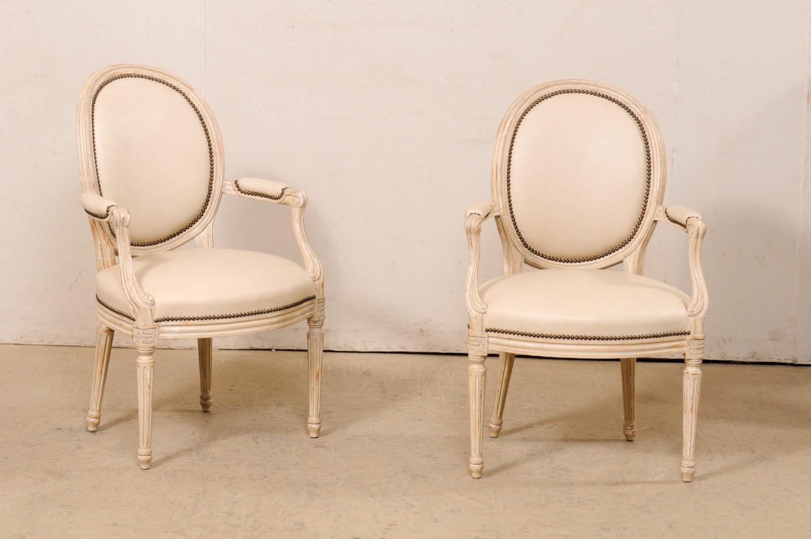 A French pair of carved/painted wood and upholstered armchairs. These vintage chairs from France each feature oval-shaped upholstered backs within a nicely molded wood frame. Manchette padded arm rests with carved curled knuckles. The skirt is nice