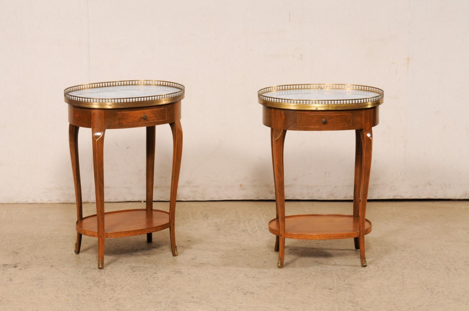 A French pair of marble and brass gallery top end tables from the mid 20th century. These vintage tables from France feature oval-shaped tops with a brass gallery edging and inset marble, upon a smooth and oval-shaped wooden apron that houses a