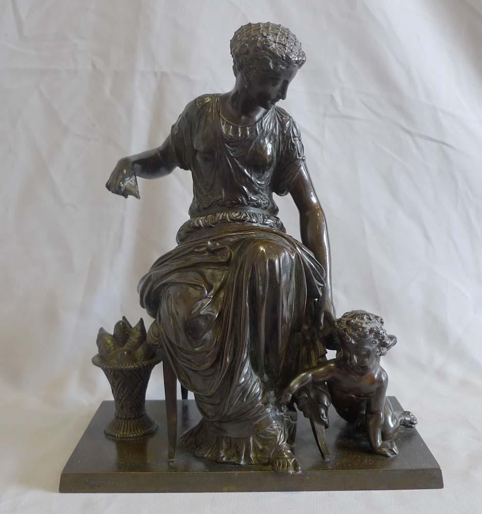 An antique French patinated bronze group of a classical woman seated on a chair and spinning thread. She has a basket of spun thread to her side. Holding a thread at her feet is the kneeling figure of a winged Cupid. The bronze is very well modelled
