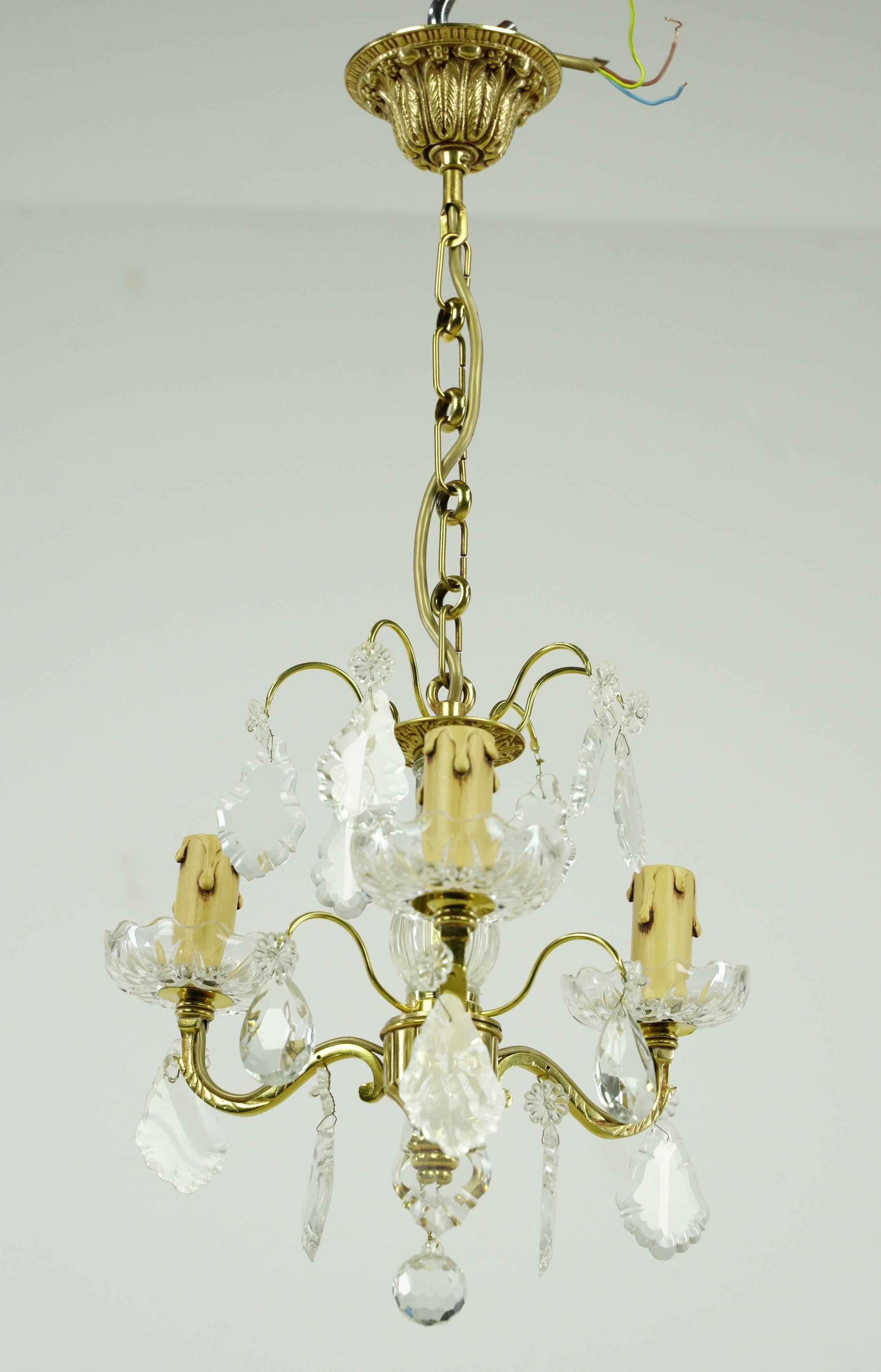 This chandelier features an ornate brass frame intricately designed to complement its petite size, adorned with clear crystals that contribute to its refined and timeless allure. This enchanting piece was sourced from an esteemed estate in