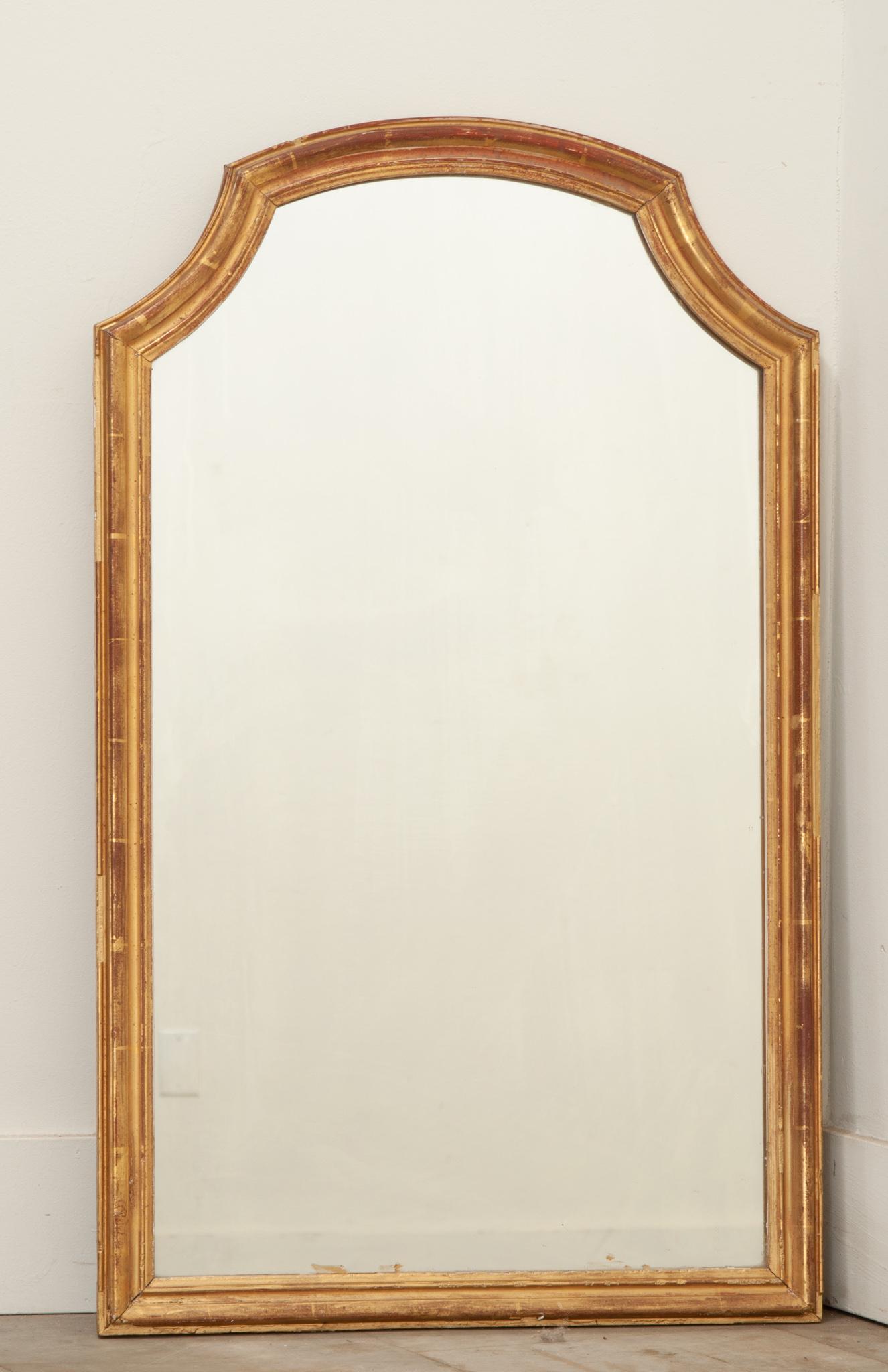 Petit miroir en bois doré fabriqué en France. La plaque de miroir est entourée d'un simple cadre doré avec un arc convexe au sommet. La dorure d'origine est patinée, révélant le bole rougeâtre en dessous, ce qui crée un contraste avec la finition