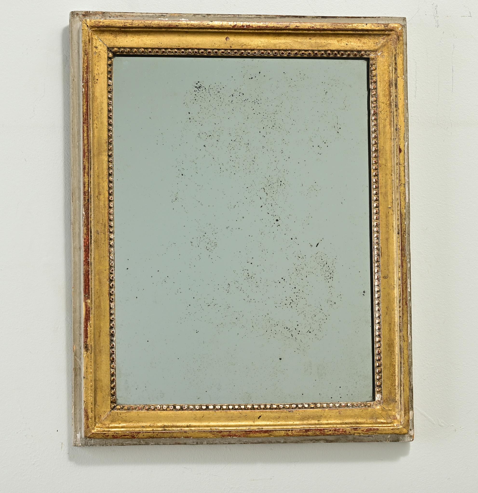 A petite French gold gilt mirror made in the Louis XVI period. This hand carved frame has its original gold gilt finish revealing the reddish bole beneath, with interior beading and a painted wood border. The symmetrical frame holds its original
