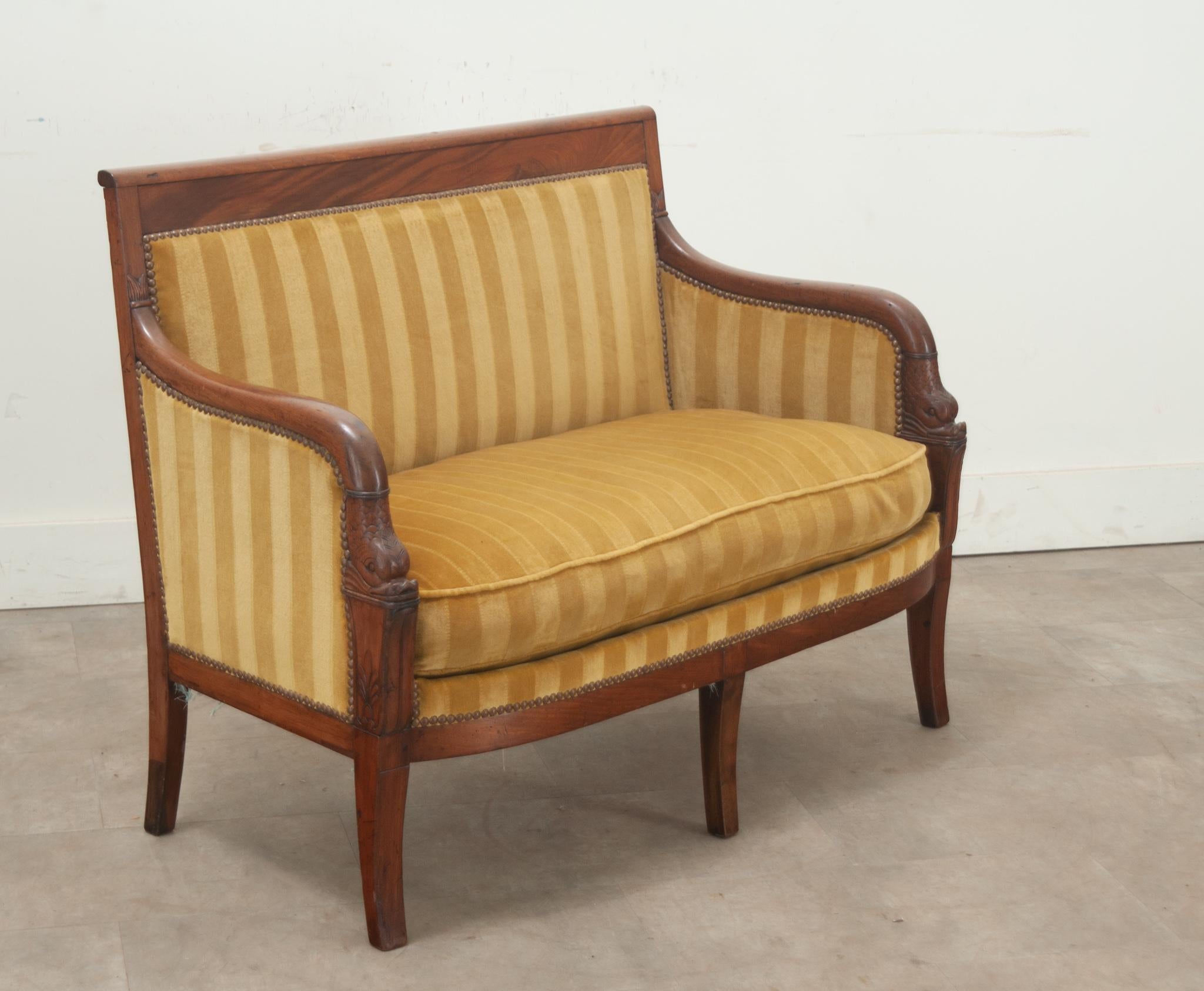A petite carved Restauration period settee made in France. This comfortable settee has a decoratively carved mahogany frame with classic motifs along the arms. The frame is upholstered in a gently used canary yellow stripe upholstery with a brass