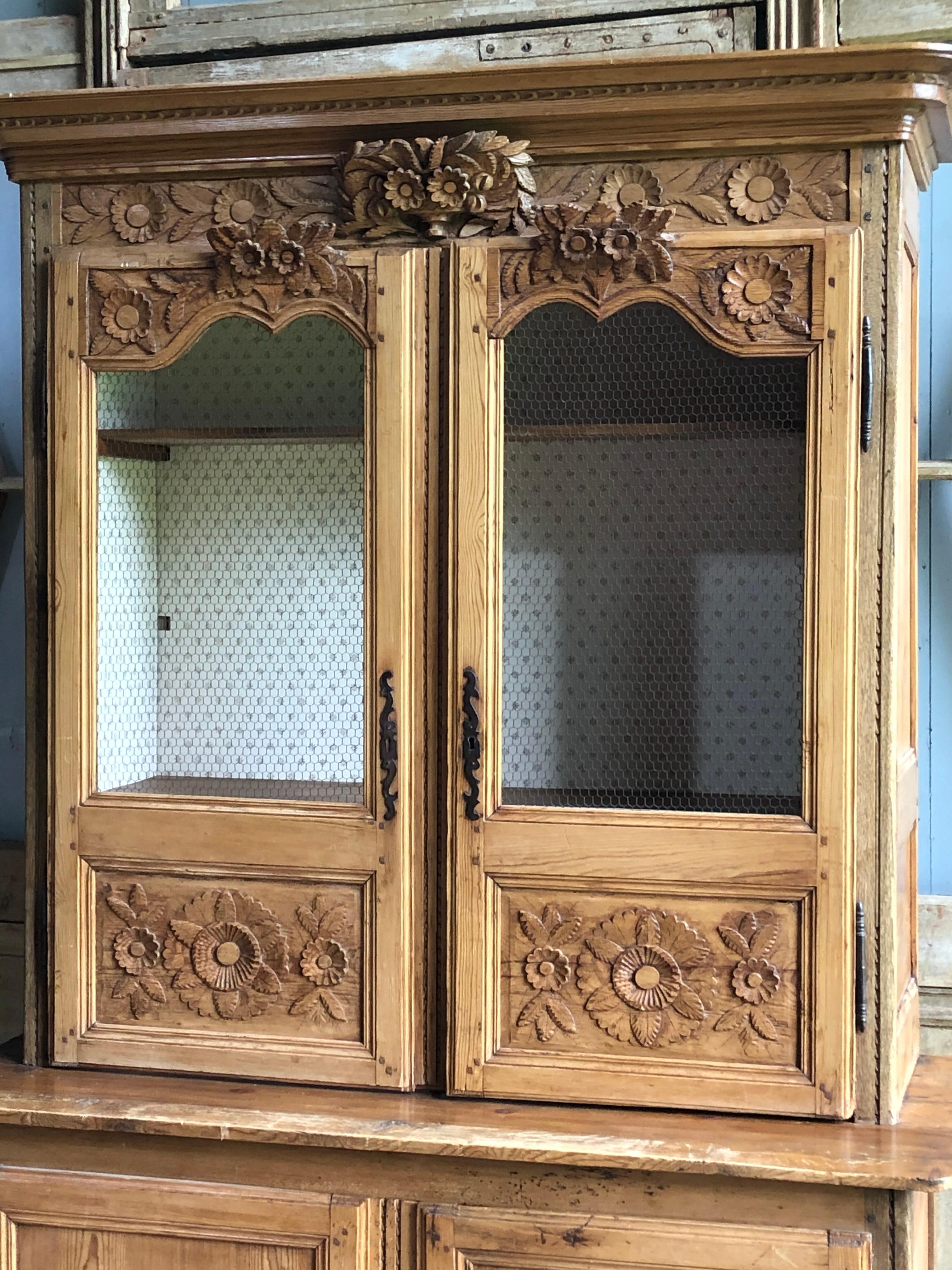 A nicely carved French buffet a deux corps in pine, from the Normandy region circa 1800, carved with floral motifs, wire grill doors in the upper cabinet and the interior lined with wallpaper. The lower cabinet was recently fitted with a drawer and
