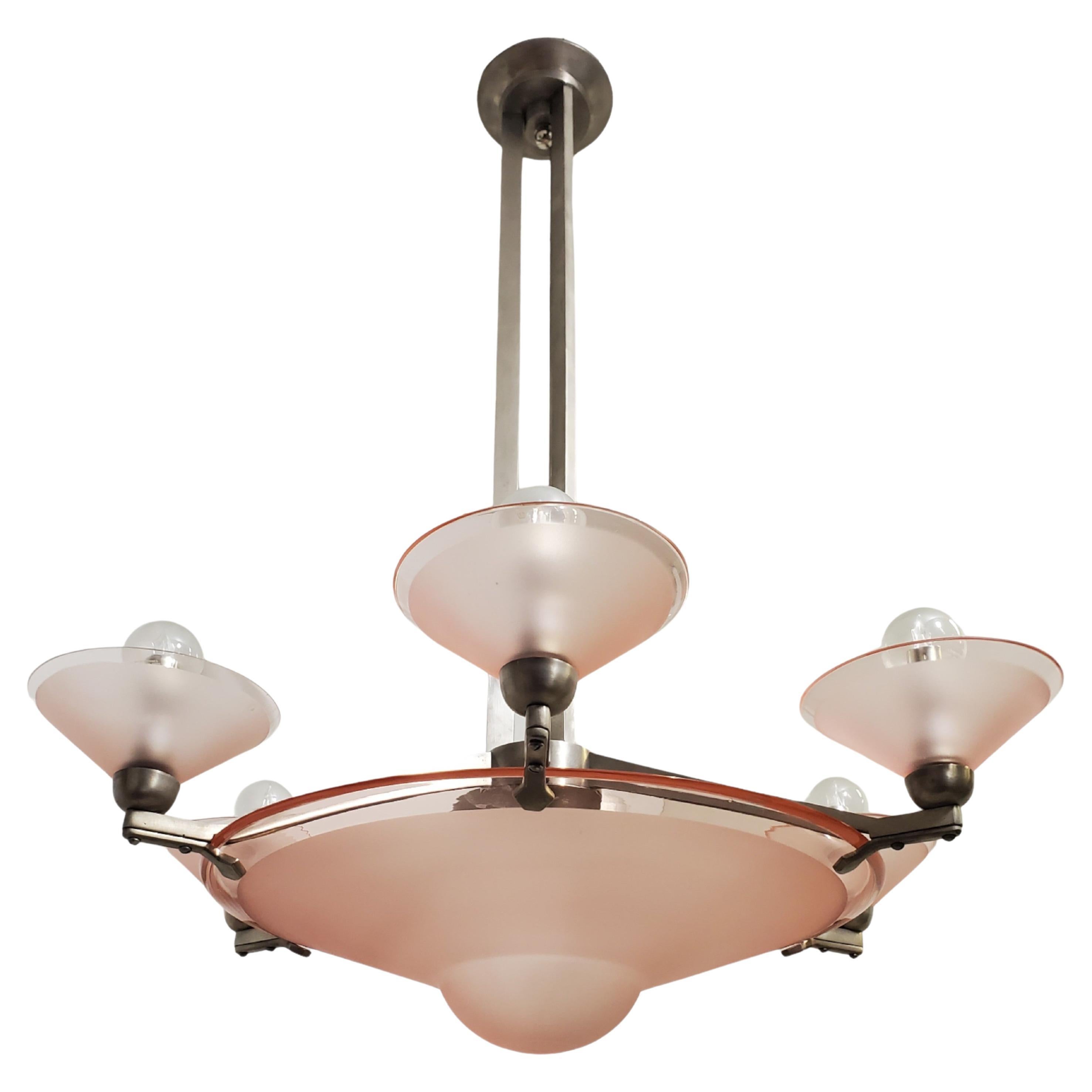 A pink frosted art glass chandelier showcasing a sizable, simple and sinuous central bowl-shaped coupe, complemented by six matching satellite dishes encircling the center. 
The chandelier is elegantly mounted on a nickeled bronze armature,