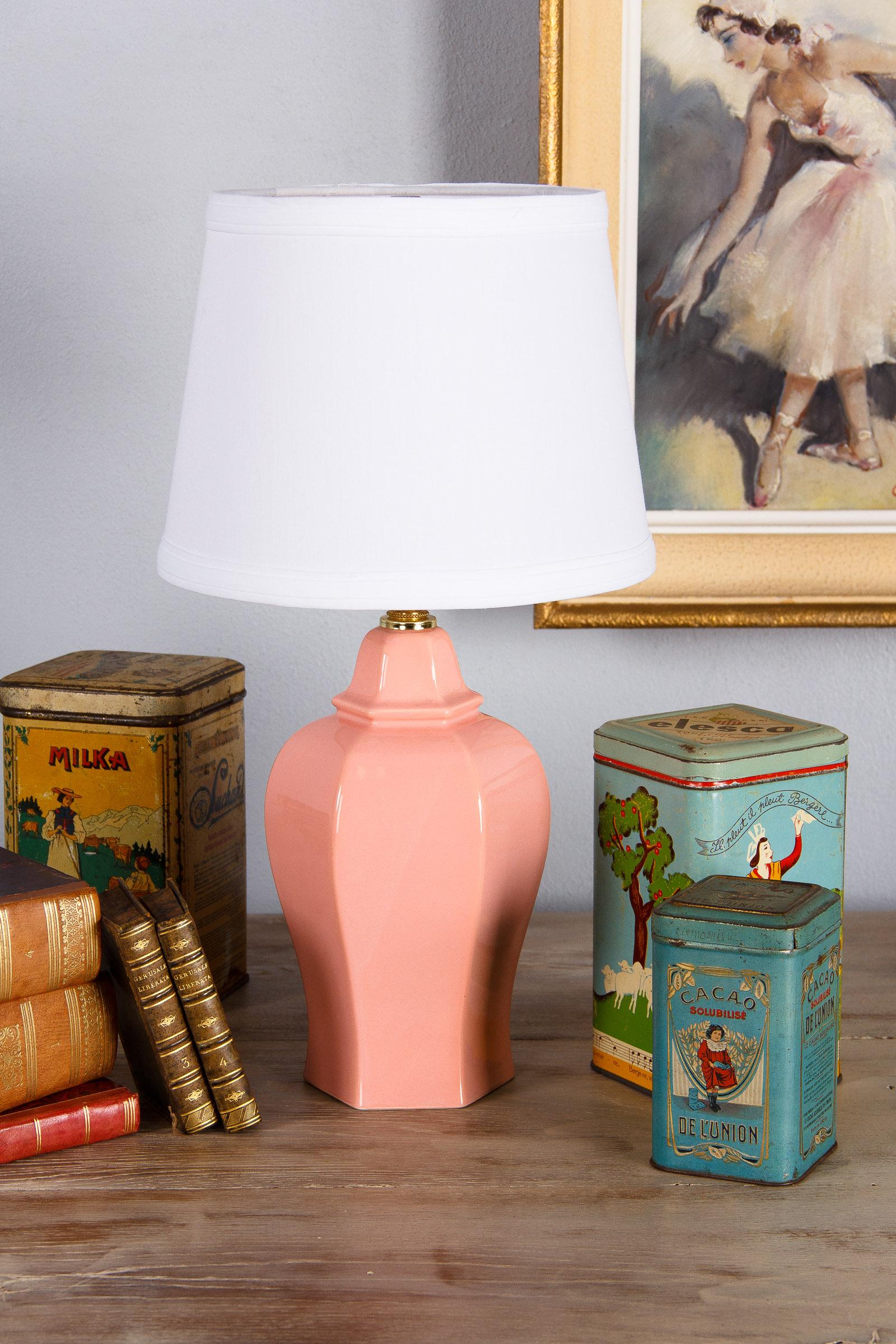 A vintage ceramic table lamp glazed in kitsch pink with white linen shade, French, circa 1960s. The six sided hollow ceramic base is shapely with a tapered bottom and wide shoulders. A brass fixture houses the three way bulb. The white linen shade