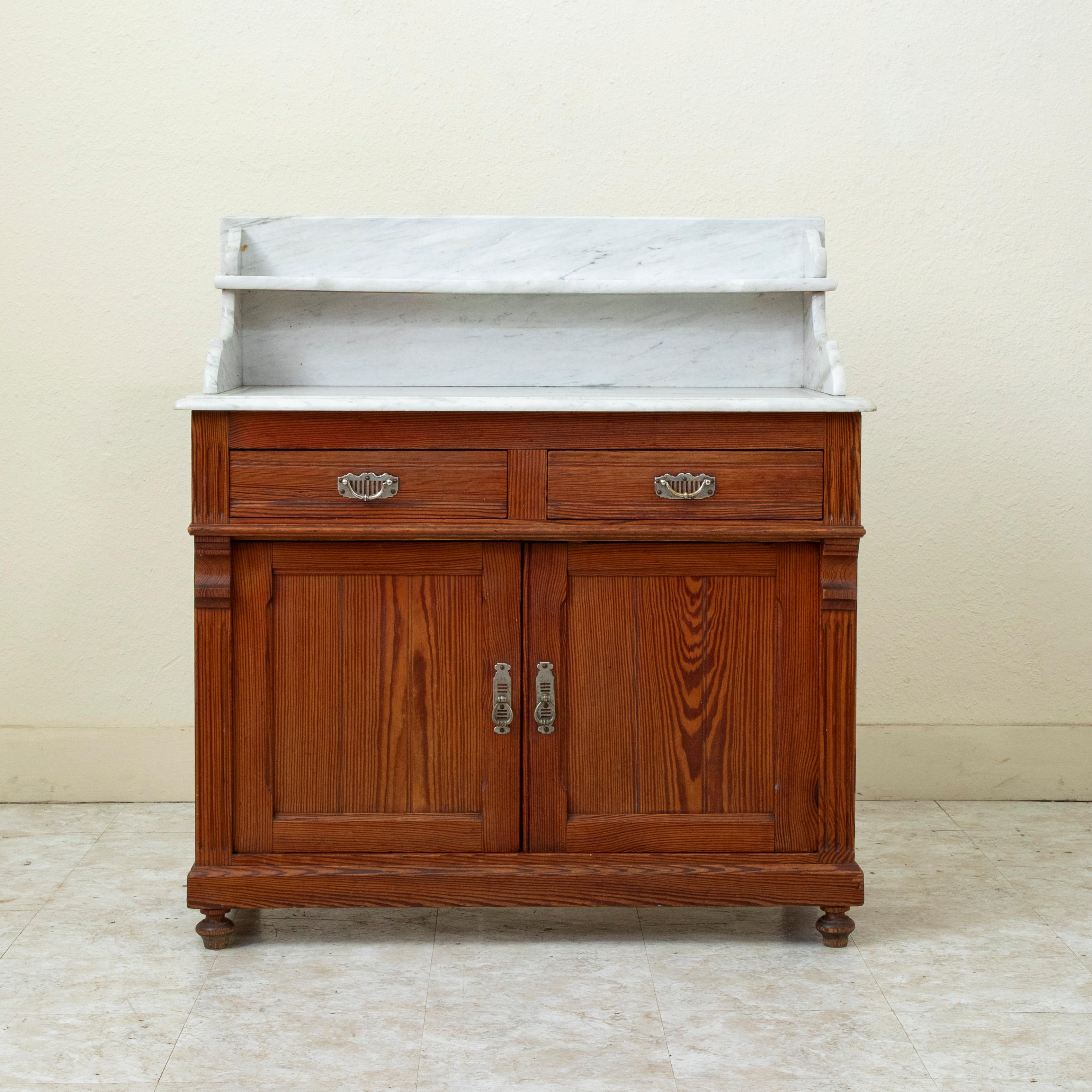 Ce meuble-lavabo en pitch pin du début du XXe siècle présente un plateau en marbre blanc biseauté avec une étagère supérieure. Ses deux tiroirs avec tirettes en métal sont construits en queue d'aronde. Sous les tiroirs se trouvent deux portes à