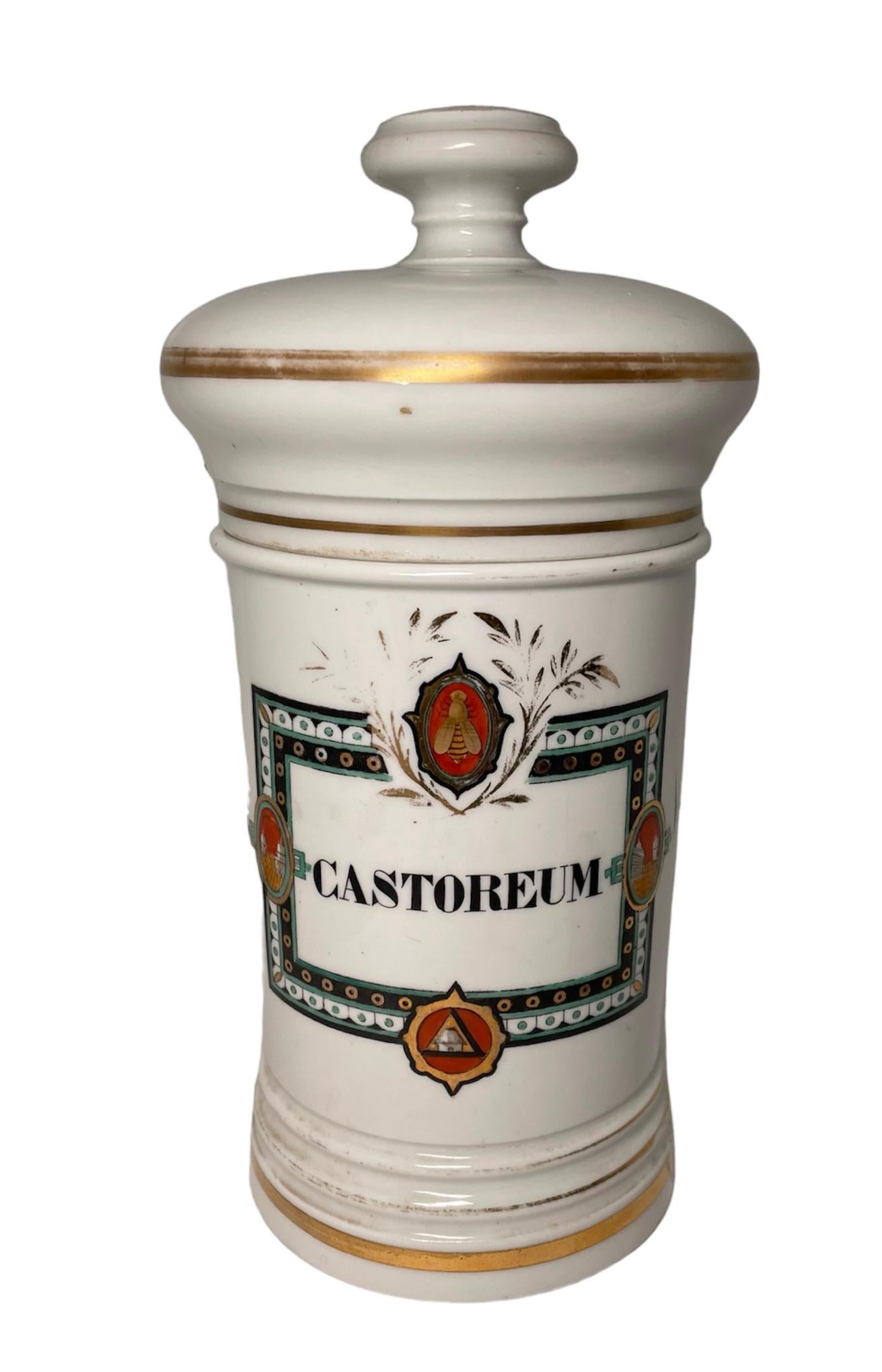 This is a French Porcelain Apothecary Jar. It depicts a cylindrical lidded jar with white background hand painted in the front with a rectangular plaque with gold borders and the name in Latin, Castoreum. The borders of the plaque are hand painted