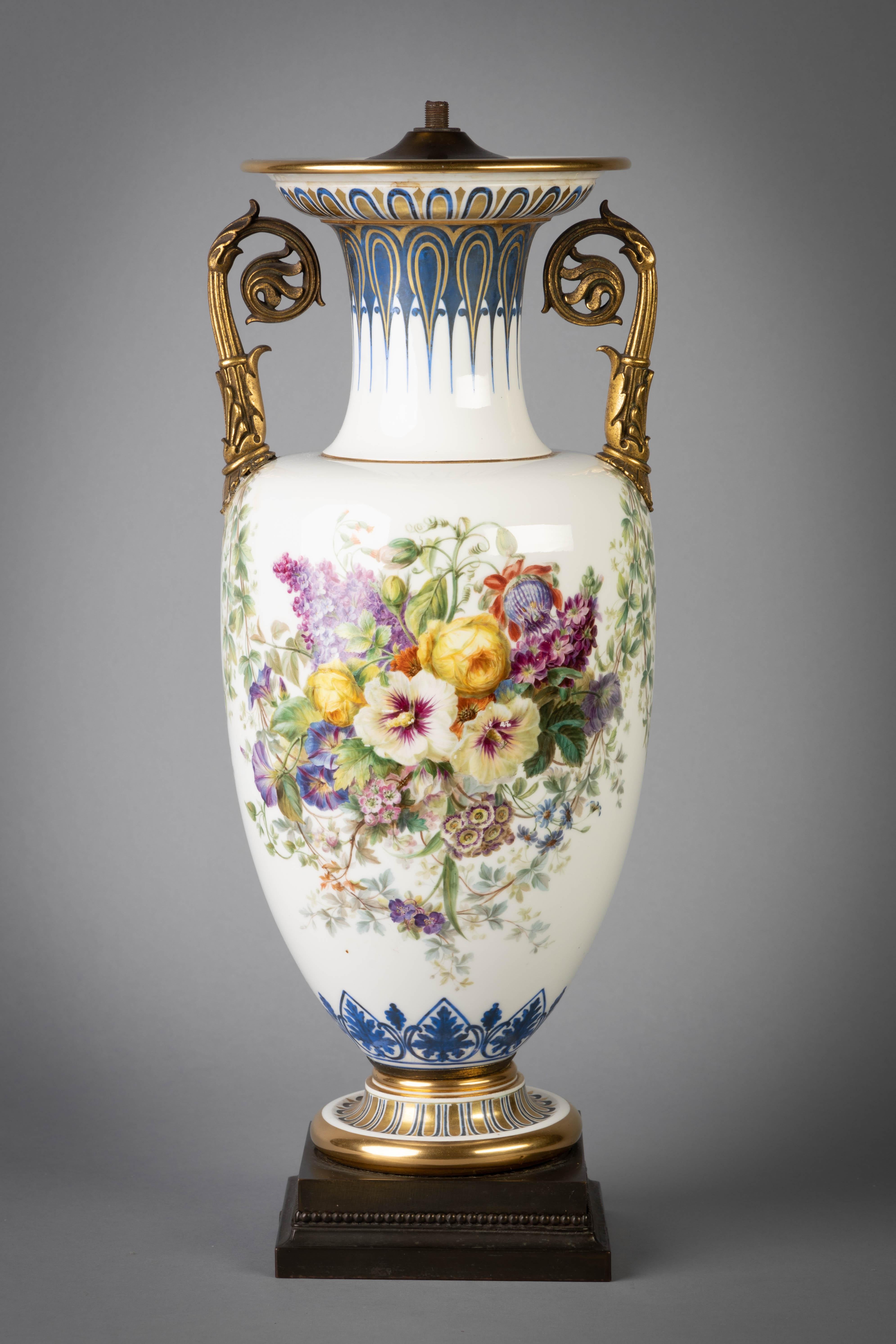 Of slender baluster form, painted with large sprays of mixed flowers hanging from vine garlands above a blue enamel lambrequin band at the base, the shoulder affixed with two gilt-bronze foliate scroll handles, with blue enamel and gilded formal