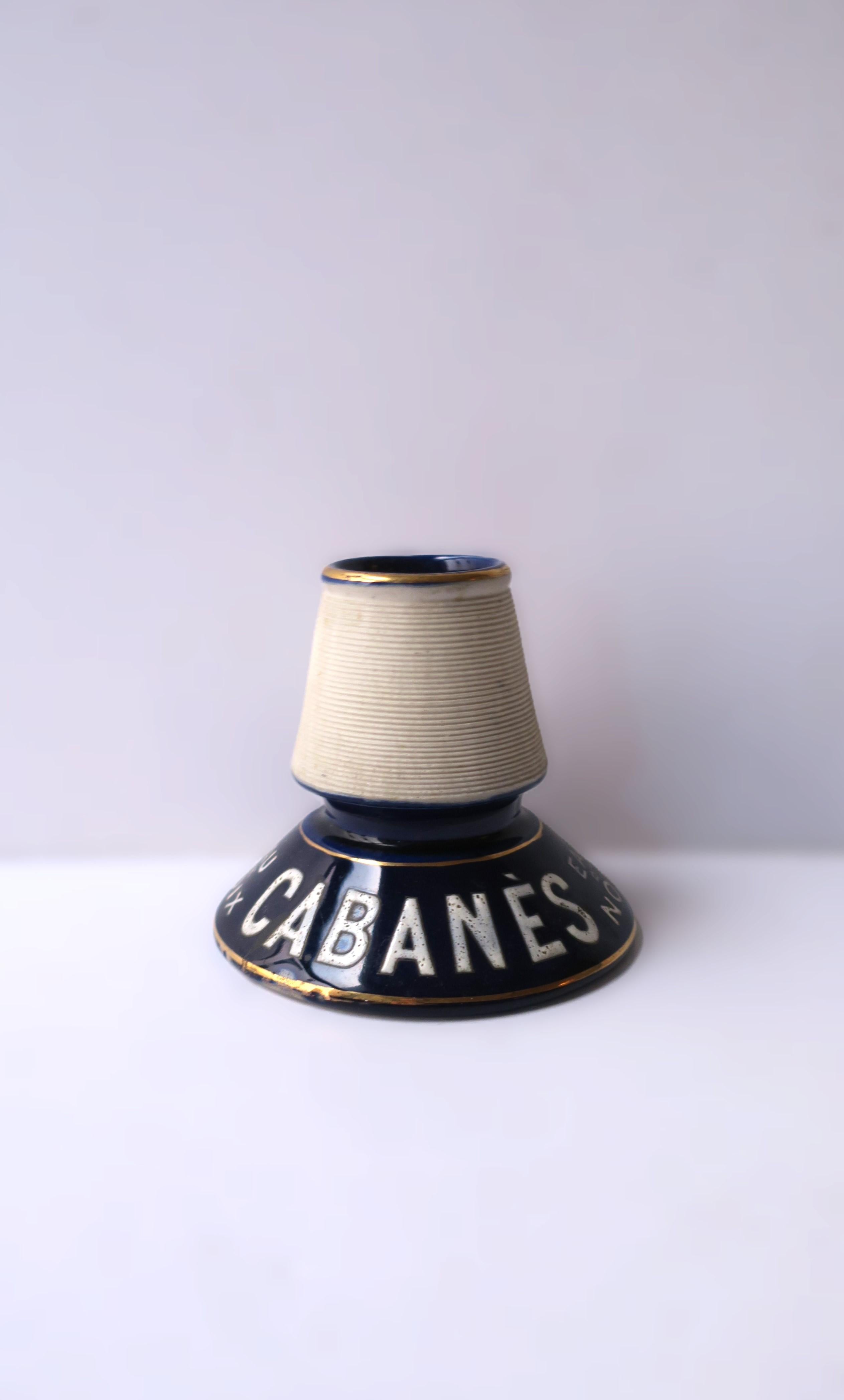 A French porcelain match striker, circa early-20th century, France. Reads: 'EAU DE NOIX CABANÉS.' Colors include a very dark blue, off-white and gold. No chips noted. 