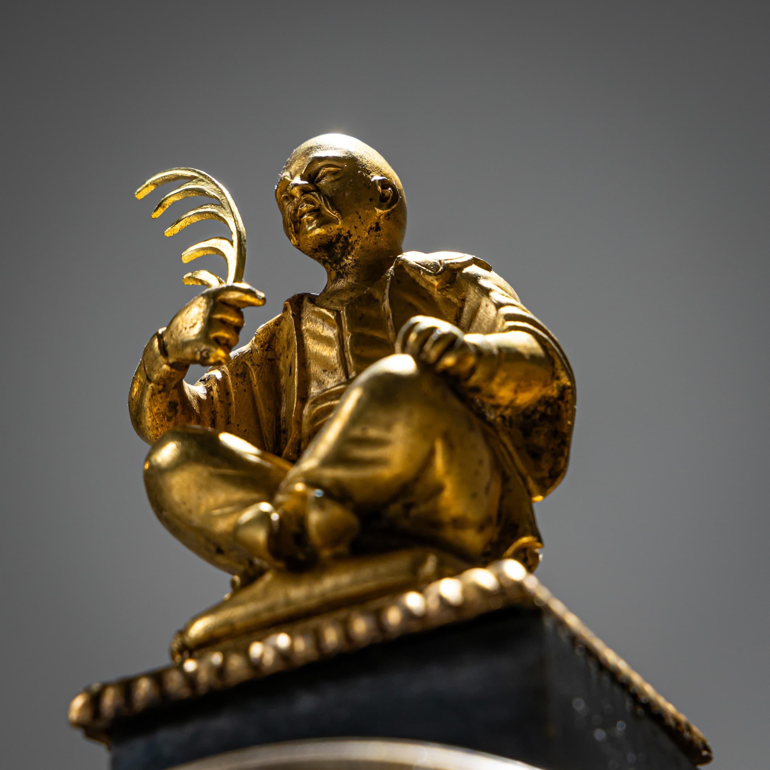 Small portico clock made of gray marble with gilded applications. The cylindrical movement is enclosed between two columns, above which a figural top piece in the form of a seated Chinese man presides. The white enameled dial with black Arabic