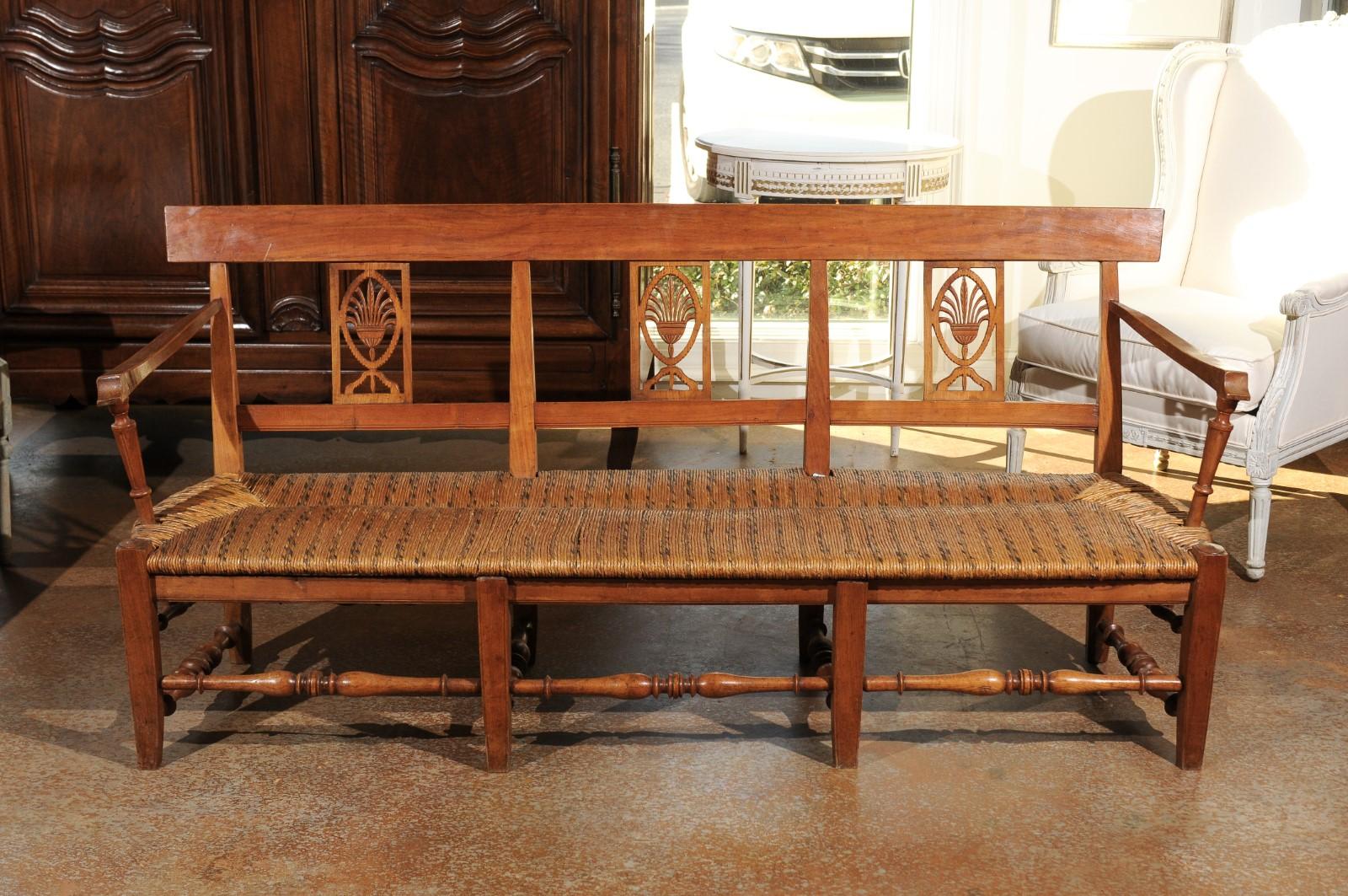 A French Provençal Directoire style wooden bench from the early 19th century, with rush seat and hand carved palmette motifs. Born in Provence during the early years of the 19th century, this exquisite French wooden bench presents the