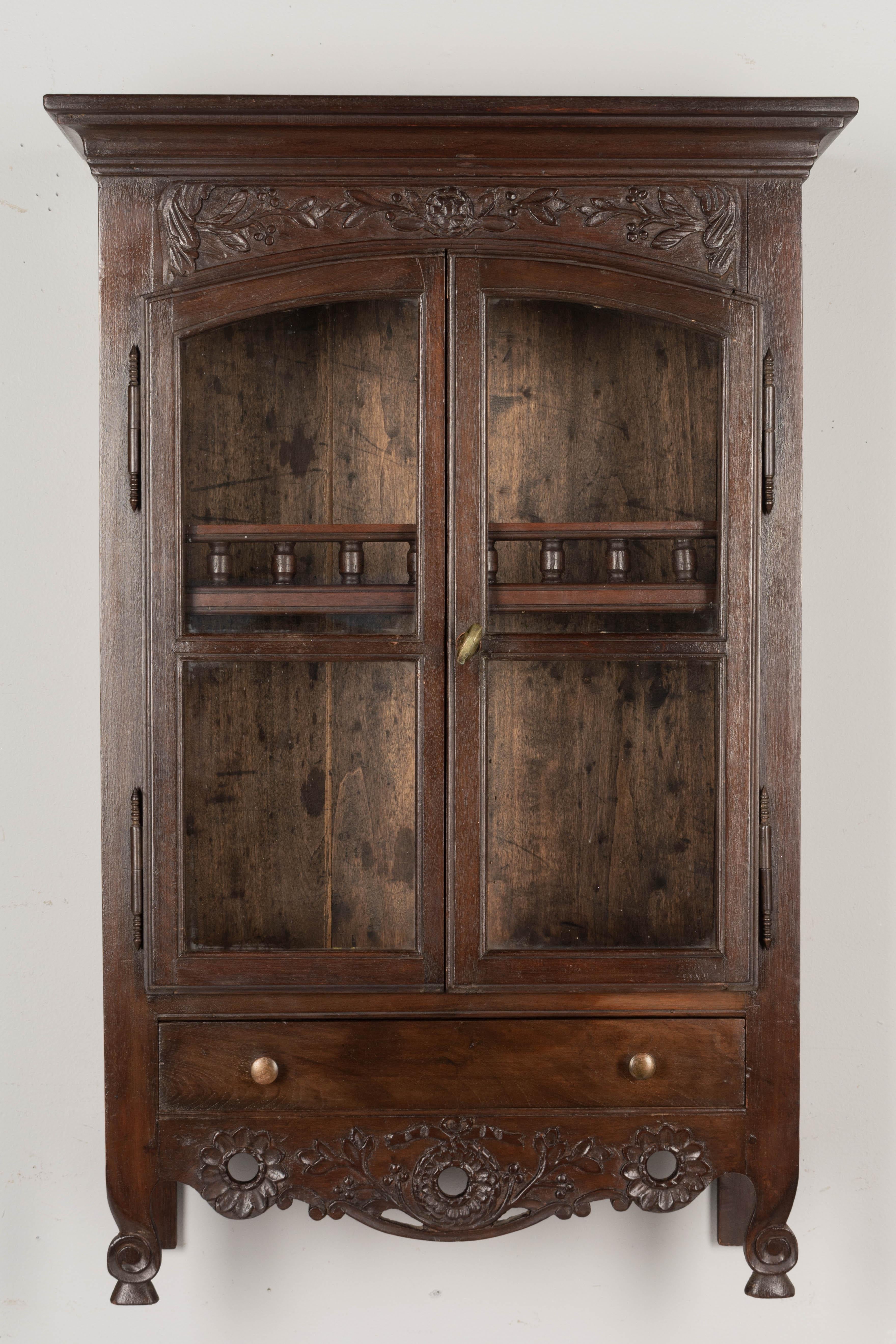A Louis XV style French Provençal vitrine, or wall display cabinet made of solid walnut with glass paned doors and a dovetailed drawer. Interior has one shelf with decorative spindle gallery. Hand-carved details at the top and a pierced carved