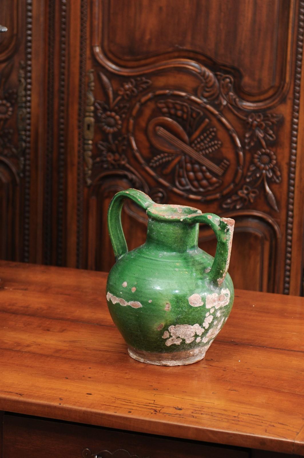 A French provincial green glazed pottery jug from the 19th century, with double handles, front spout and weathered patina. Created in Southern France during the 19th century, this jug features a green glazed body showcasing a nicely distressed