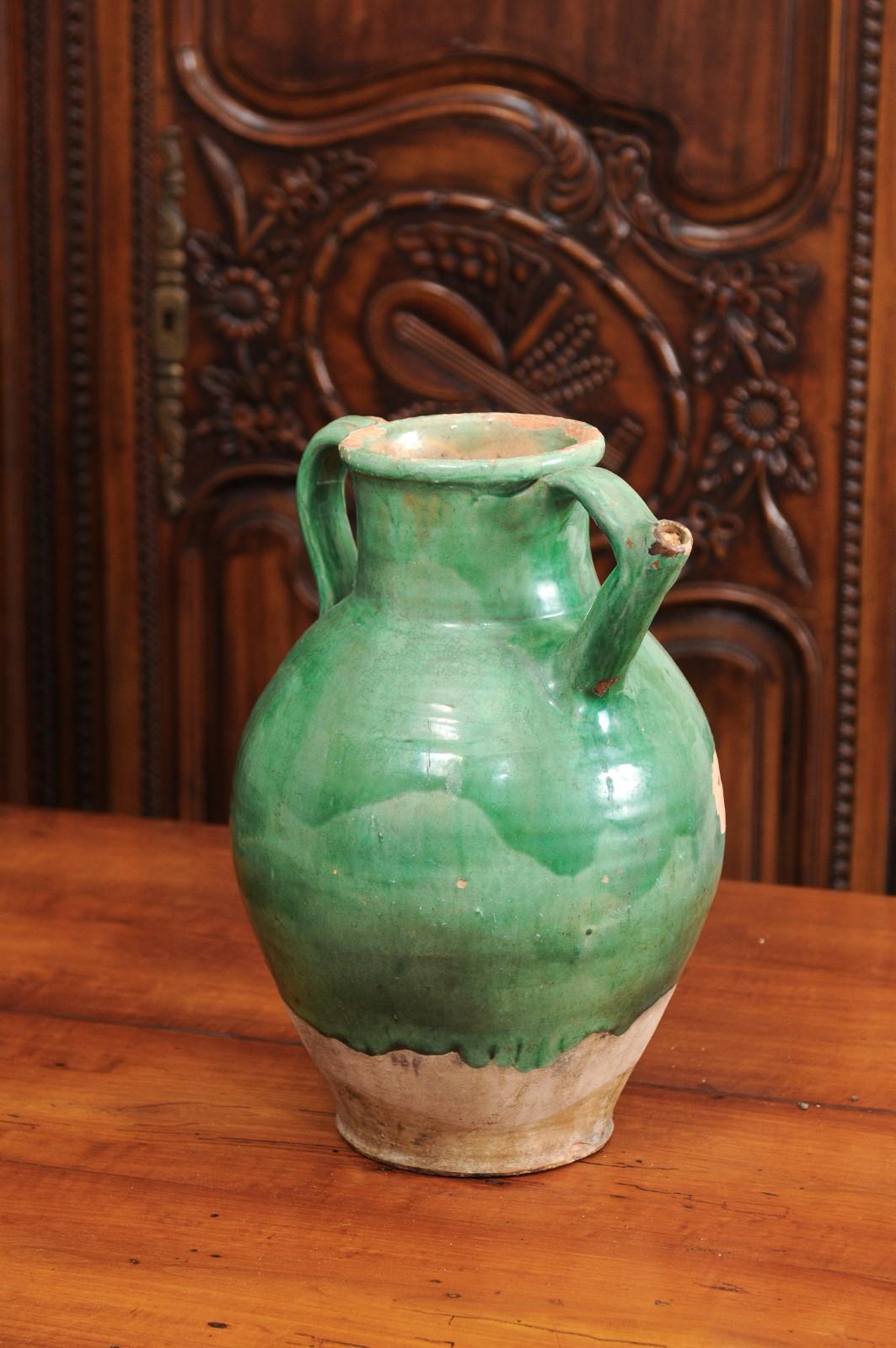 A French provincial green glazed pottery jug from the 19th century, with double handles, front spout and unfinished base. Created in France during the 19th century, this jug features a green glazed body showcasing a nicely distressed patina.