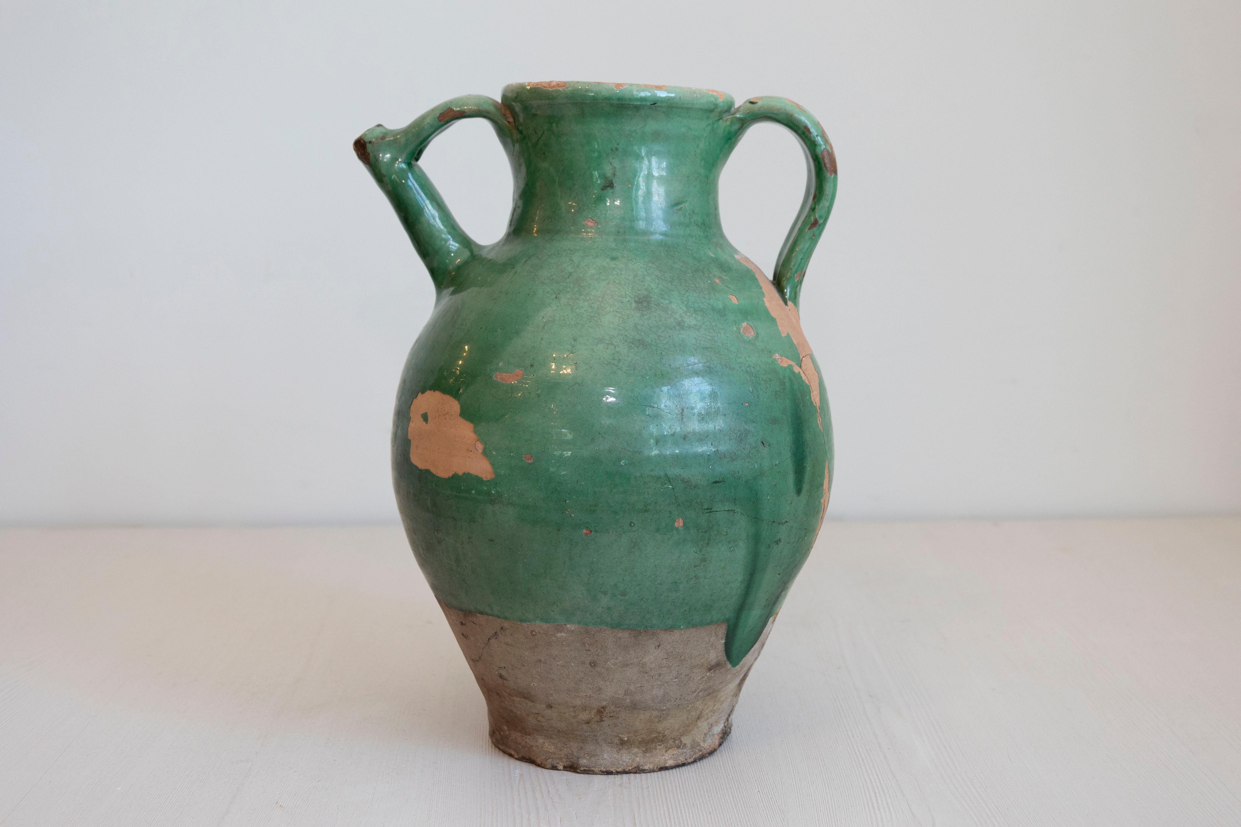 Jarra de cerámica vidriada verde provincial francesa del siglo XIX, con asas dobles, pico frontal y base inacabada. Creada en Francia en el siglo XIX, esta jarra presenta un cuerpo esmaltado en verde con una pátina muy desgastada. Acentuada con una