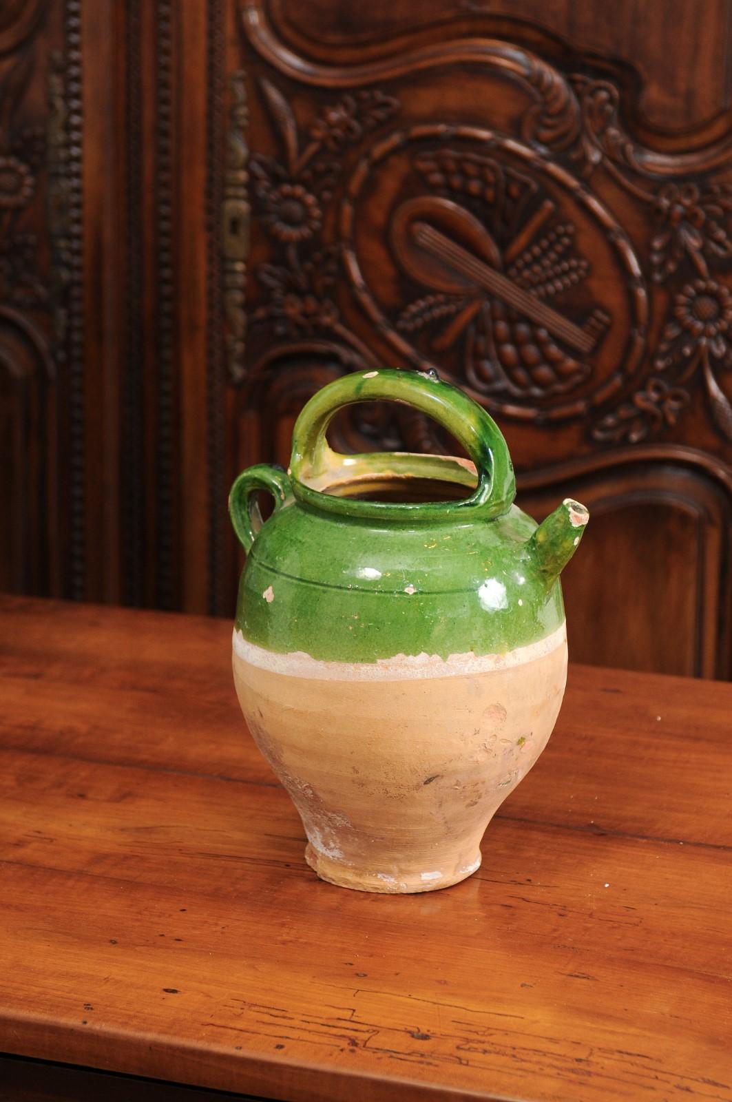 Une cruche en poterie émaillée verte de style provincial français du 19e siècle, avec deux poignées, un bec verseur frontal et une patine vieillie. Créée dans le sud de la France au cours du XIXe siècle, cette cruche présente une section supérieure