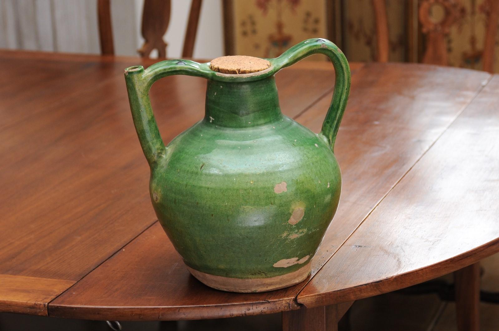 A French Provincial green glazed pottery pitcher from the 19th century, with double handle, cork top, front spout and distressed patina. Created in Southern France during the 19th century, this pitcher features a green glazed body showcasing a