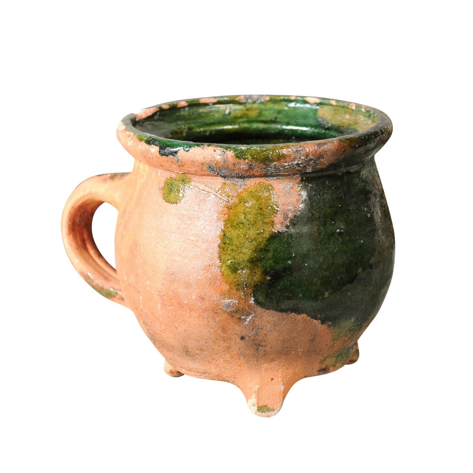 A French Provincial pottery from the 19th century with green glaze, molded handle, tripod base and weathered patina. Crafted with precision and artistry, this French Provincial pottery showcases a charming green glaze that adds a touch of elegance