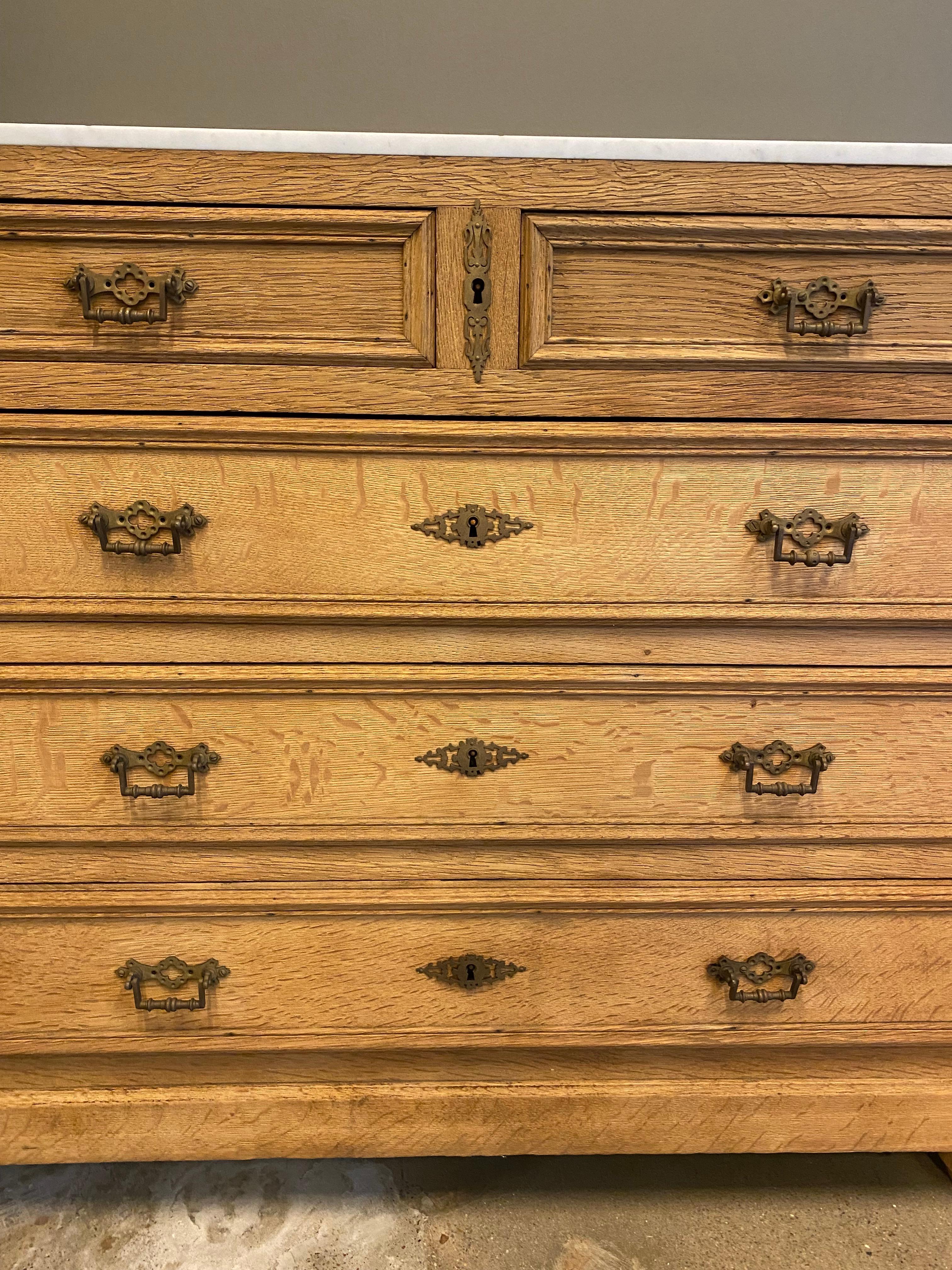 19th century country French provincial commode or chest of drawers crafted from oak with a bleached finish. The chest features a white Carrara marble-top with evidence of a prior wash stand backing. Fitted with five storage drawers the case has