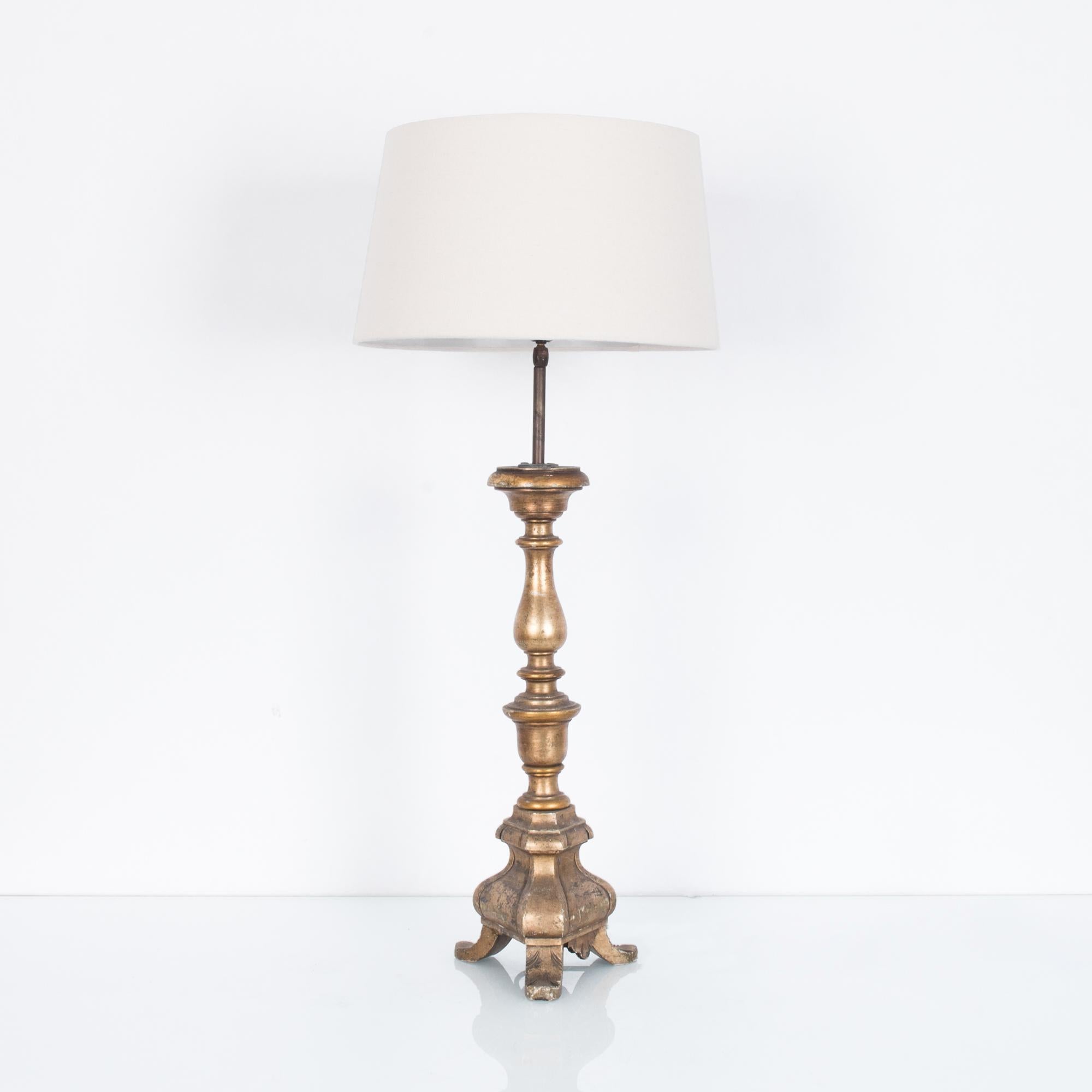 A gilded wooden table lamp from France, circa 1800. A repurposed wooden candlestick supports an empire lampshade in natural white. The shape is slender and elaborate, elevated by three curved feet. The gilded finish has aged into a warm, brassy
