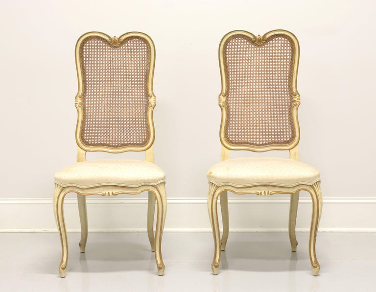 A pair of vintage French Provincial Louis XV style dining side chairs, unbranded, possibly by Davis Cabinet Co. Cherry wood painted an antique white with gold accents, cane backs, fabric upholstered seats, curved legs and scroll feet. Made in the