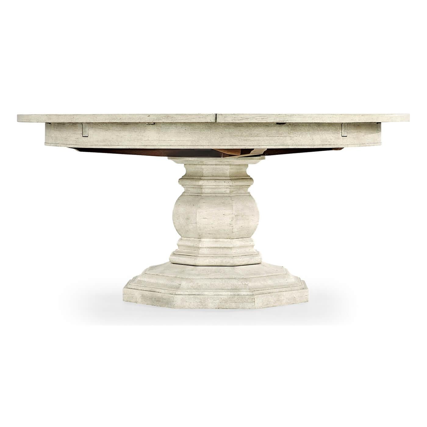 A French Provincial style whitewash driftwood topped round dining table with a rustic finish showing exposed saw marks set on a substantial baluster stem base, the table extending via an ingenious mechanism of self-storing hinged fold-out leaves